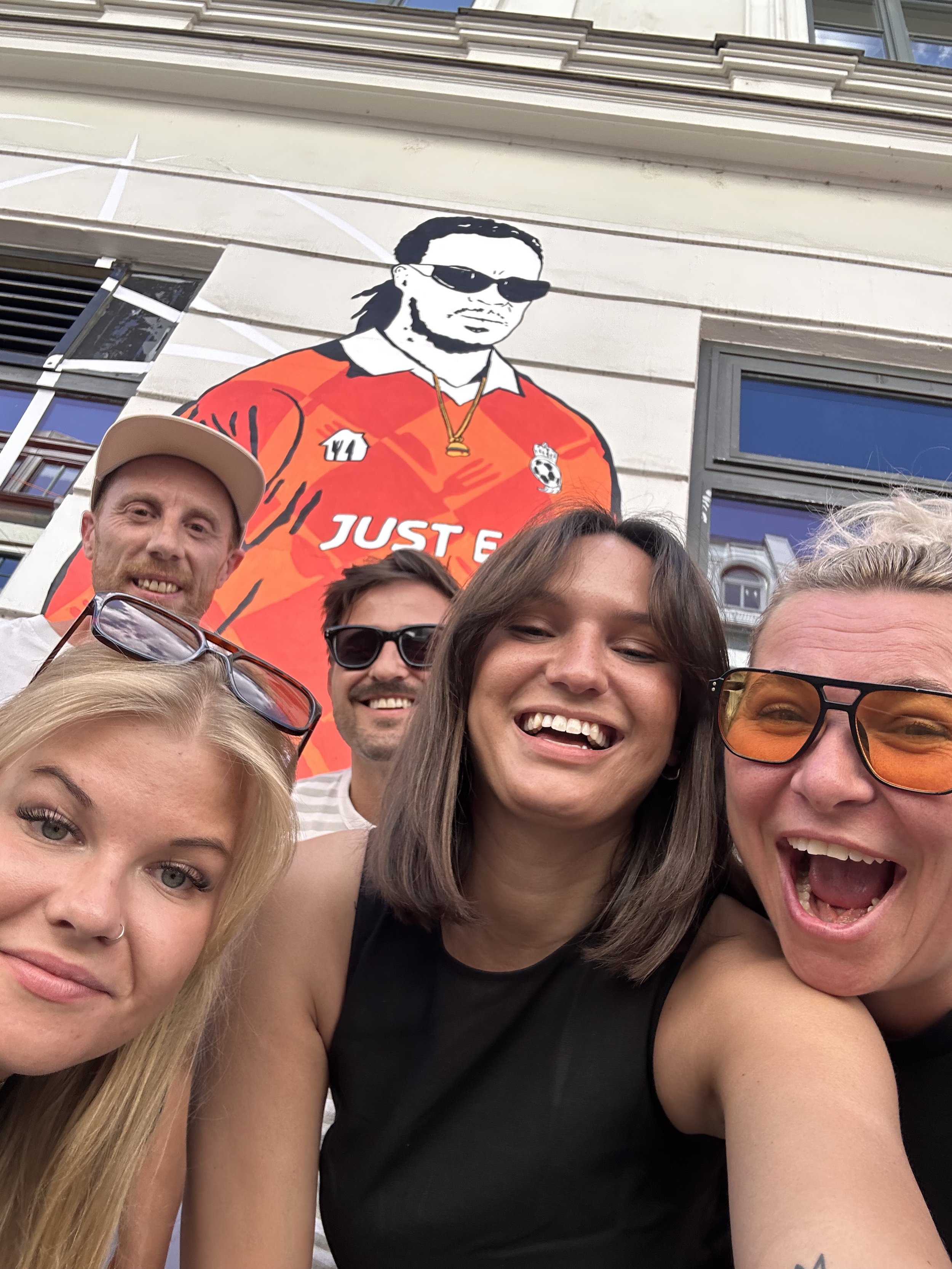 Gruppe von fünf smiling Menschen, drei Frauen und zwei Männer, die ein Selfie vor einem Wandbild von einem Mann in Sonnenbrille auf einer Häuserwand machen. Durch Folierung und Grafikdesign erstellt.