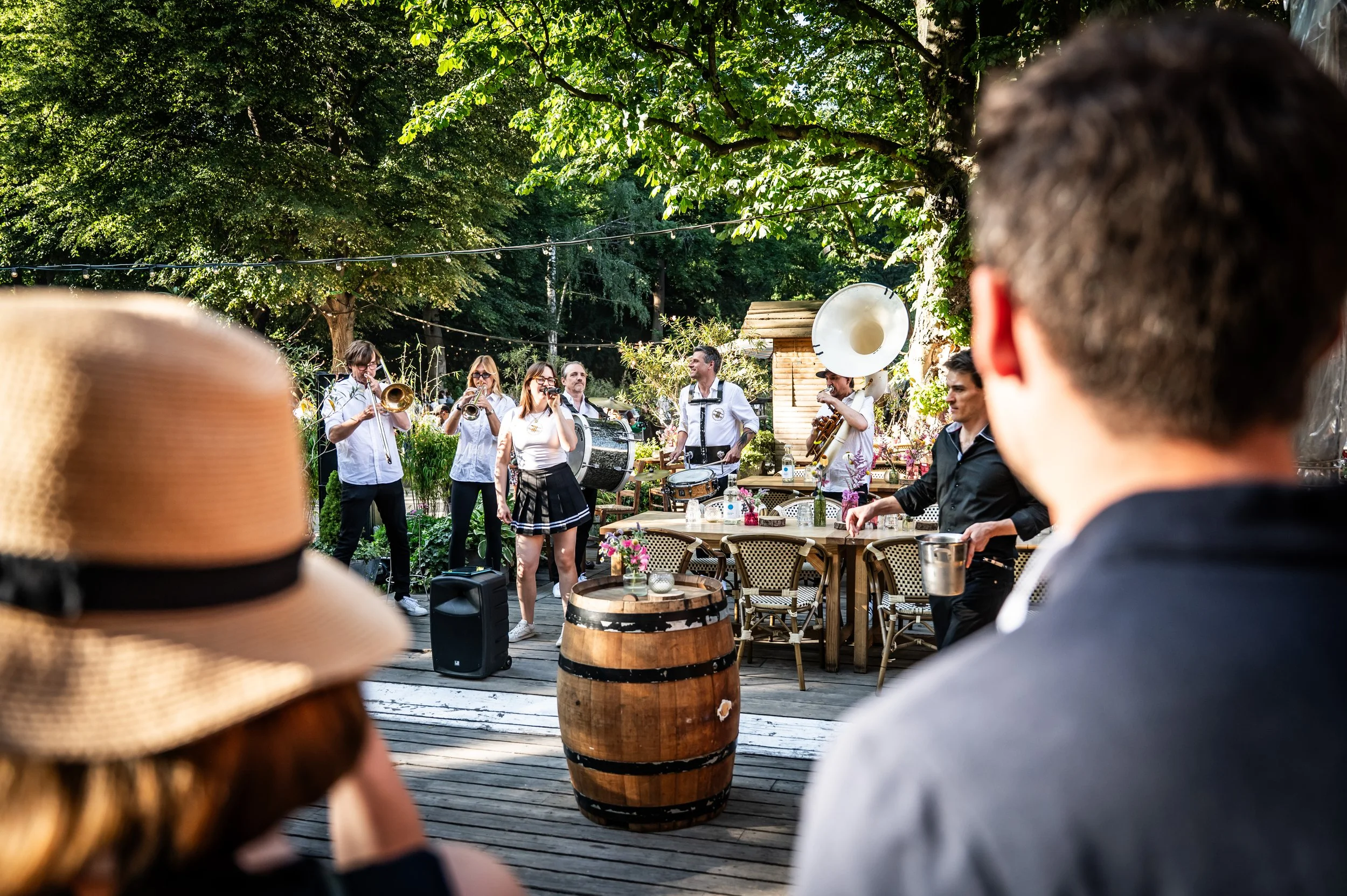 Musikkapelle spielt live im Freien. Zuschauer, darunter eine Frau mit Strohhut, sitzen an Tischen und genießen die Aufführung. Es ist ein sonniger Tag im Garten mit vielen Bäumen.
