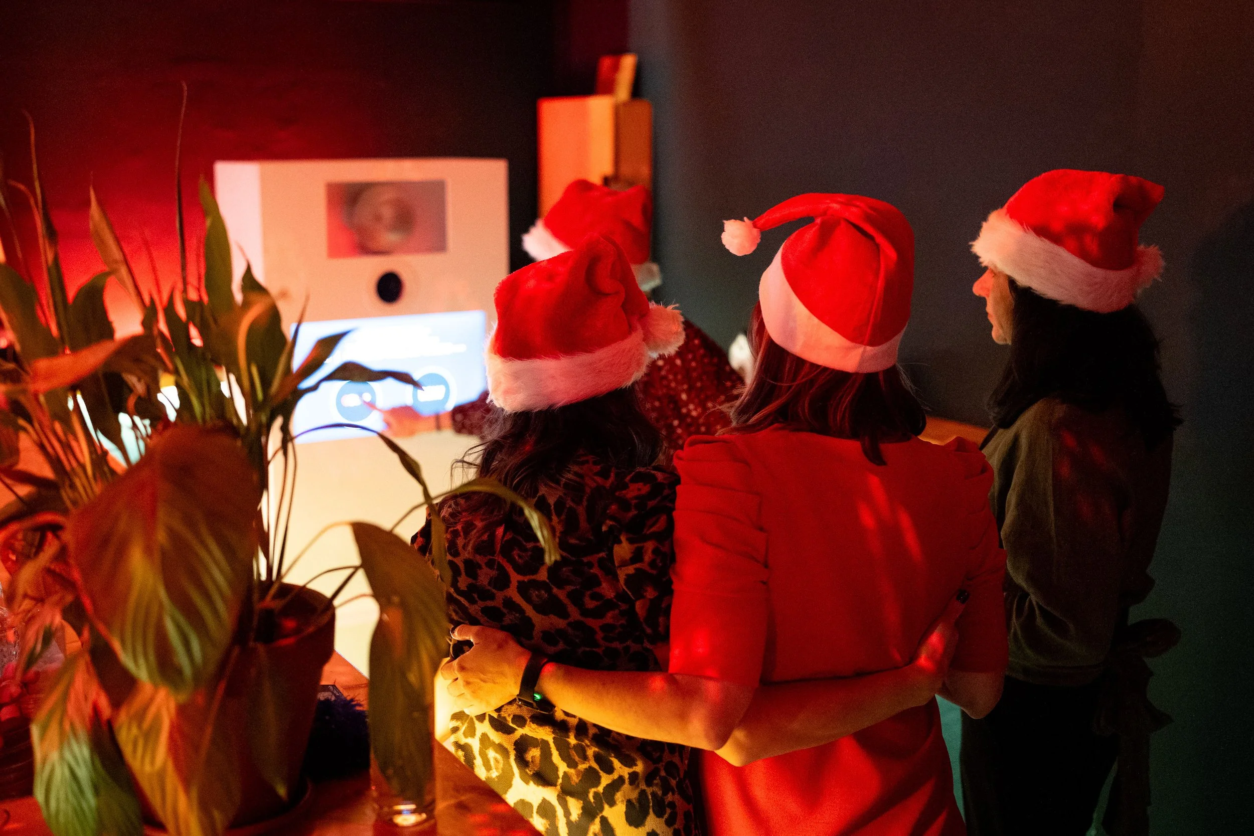 People wearing Santa hats gathered around at a holiday celebration, with a dimly lit background and festive atmosphere.