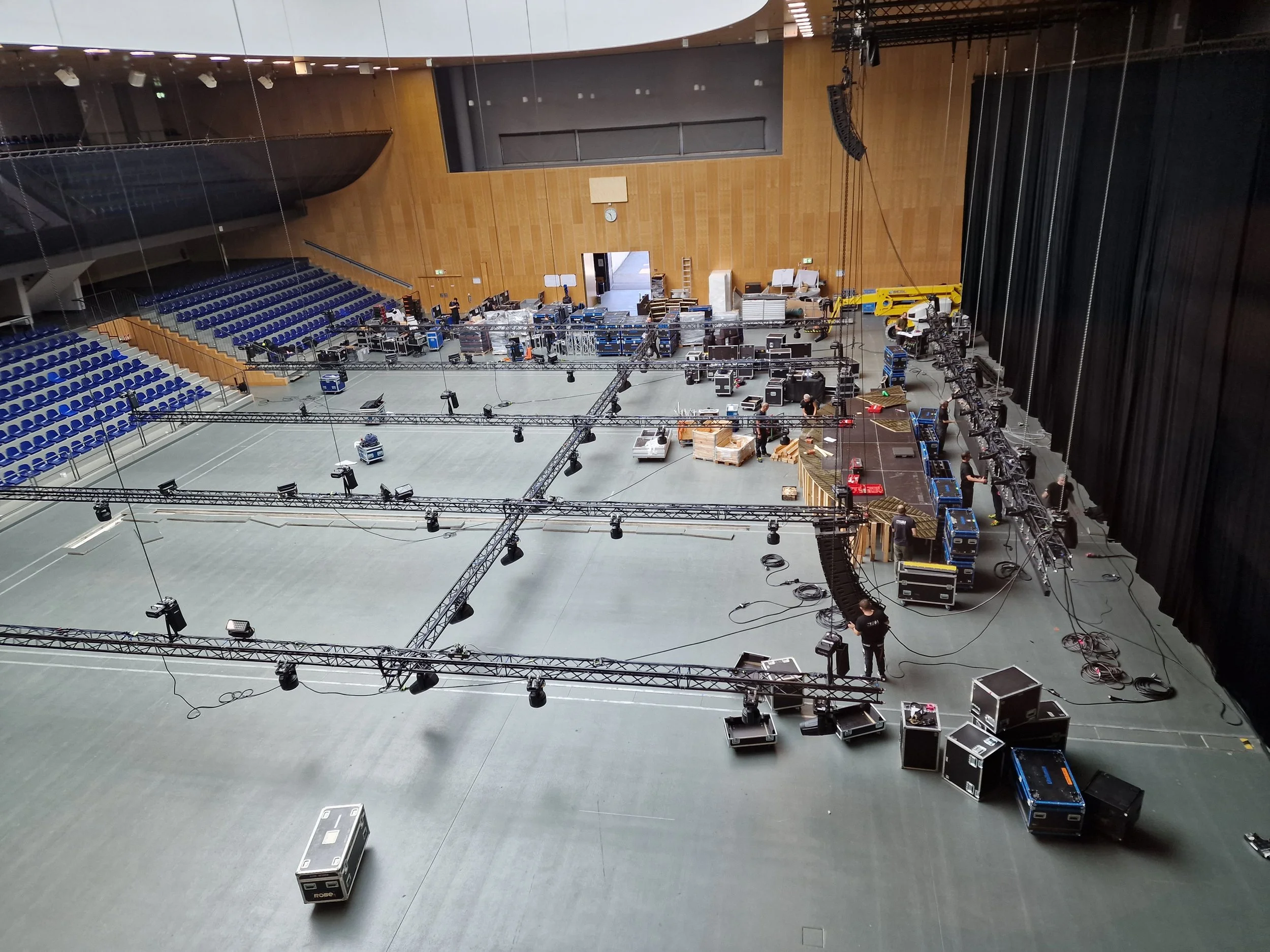 Ein Veranstaltungsraum wird für eine Bühne aufgebaut. Es gibt zahlreiche Lichter, Kabel und technische Ausrüstung. Im Hintergrund sind Sitzreihen mit blauen Stühlen. Es sind einige Personen bei der Einrichtung der Beleuchtung und Technik beteiligt.