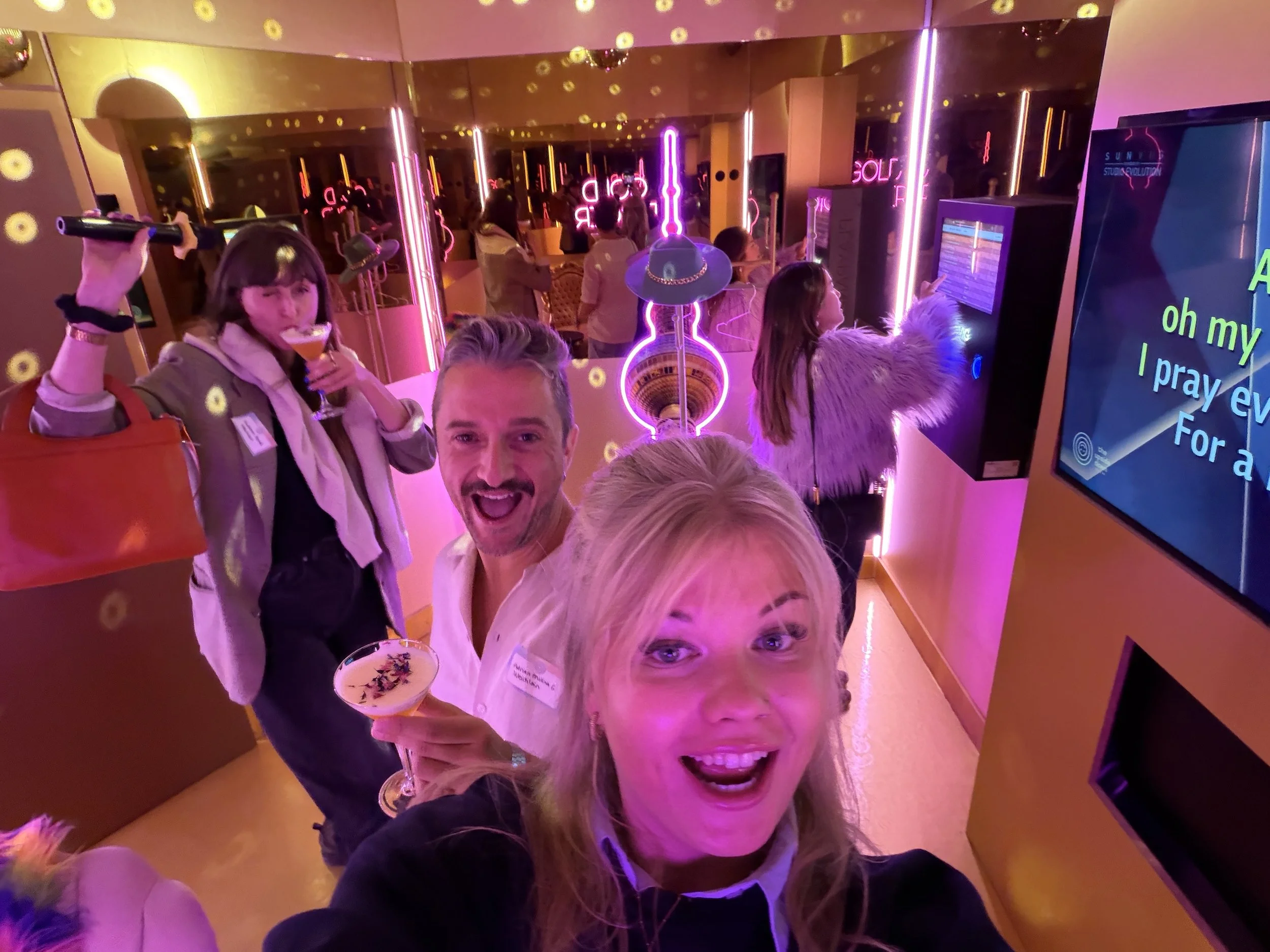 Multiple people at a lively party or nightclub with neon lighting, mirrors, and vibrant decor, engaging in conversation and enjoying drinks.