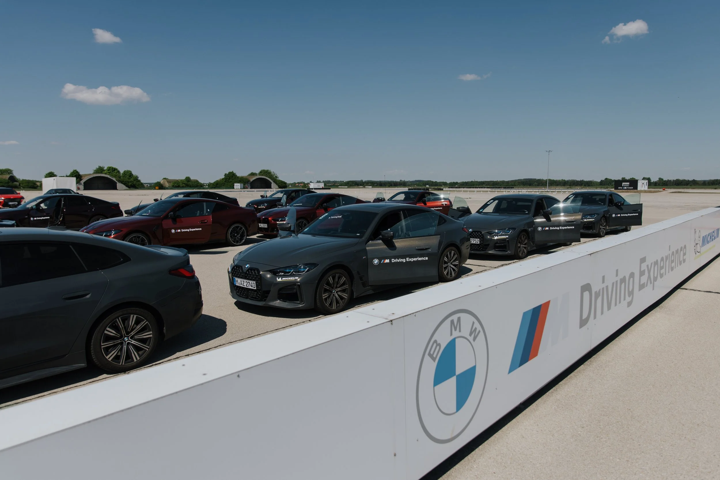 Multiple BMW and other cars parked on a driving experience track under a blue sky with a few clouds. The cars are arranged in a line behind a white barrier with BMW and other logos. The setting is open with a flat landscape and distant trees.