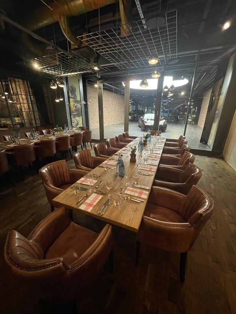 Innenraum eines eleganten Restaurants mit einem langen Holztisch, umgeben von braunen Ledersesseln. Der Tisch ist mit Gläsern, Besteck, Stoffservietten und Wasserflaschen gedeckt. Im Hintergrund sind große Glasfenster, die auf einen Parkplatz mit geparkten Autos blicken. Die Beleuchtung ist warm und modern, mit Hängelampen, die von der Decke hängen.