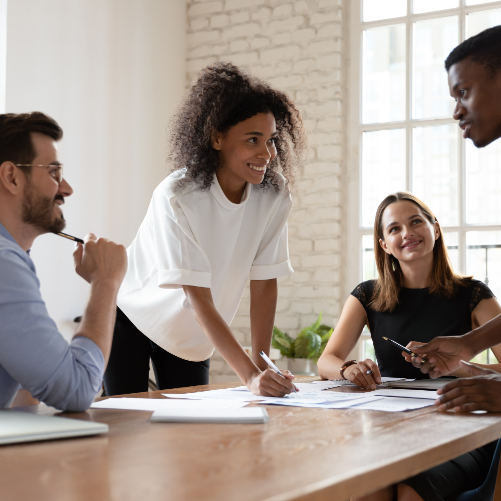 Four diverse people collaborating around a wooden table in a bright, modern office with a large window and white brick wall.