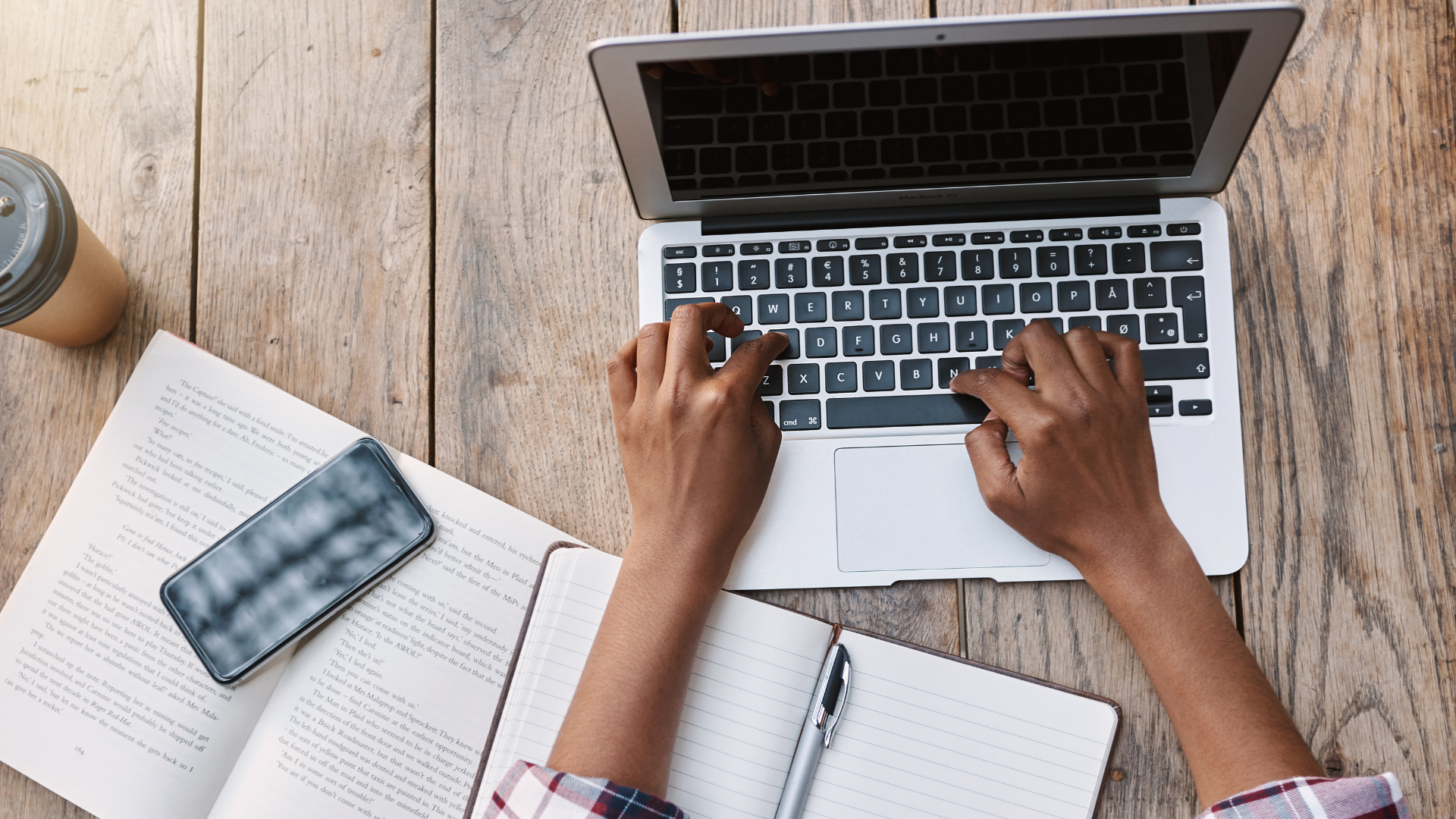 A person typing on a laptop at a wooden desk with an open book, a smartphone, a notebook with a pen, and a cold beverage.