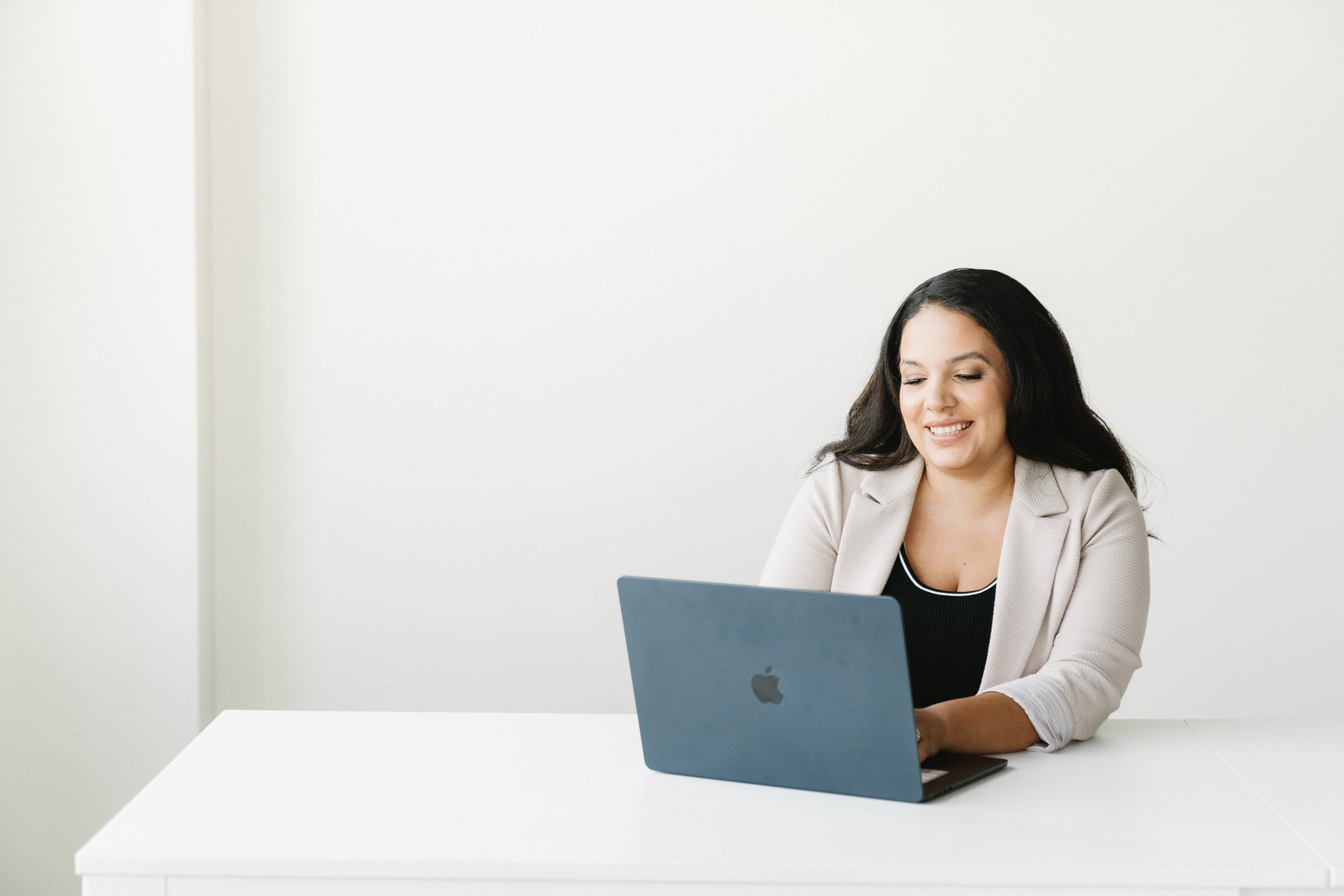 Smiling woman sitting at white desk using Apple laptop in a minimal, bright room.