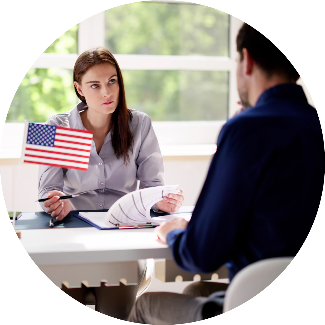 A woman holding a pen and paper, sitting at a desk with an American flag, talking to a man across from her in an office with a window showing green trees outside.
