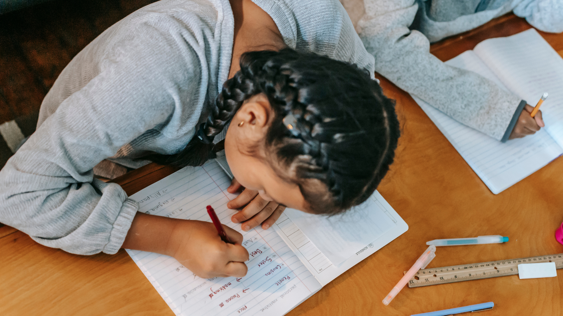 A girl with braided hair writing in a notebook on a wooden table, surrounded by pens, a ruler, and additional notebooks.
