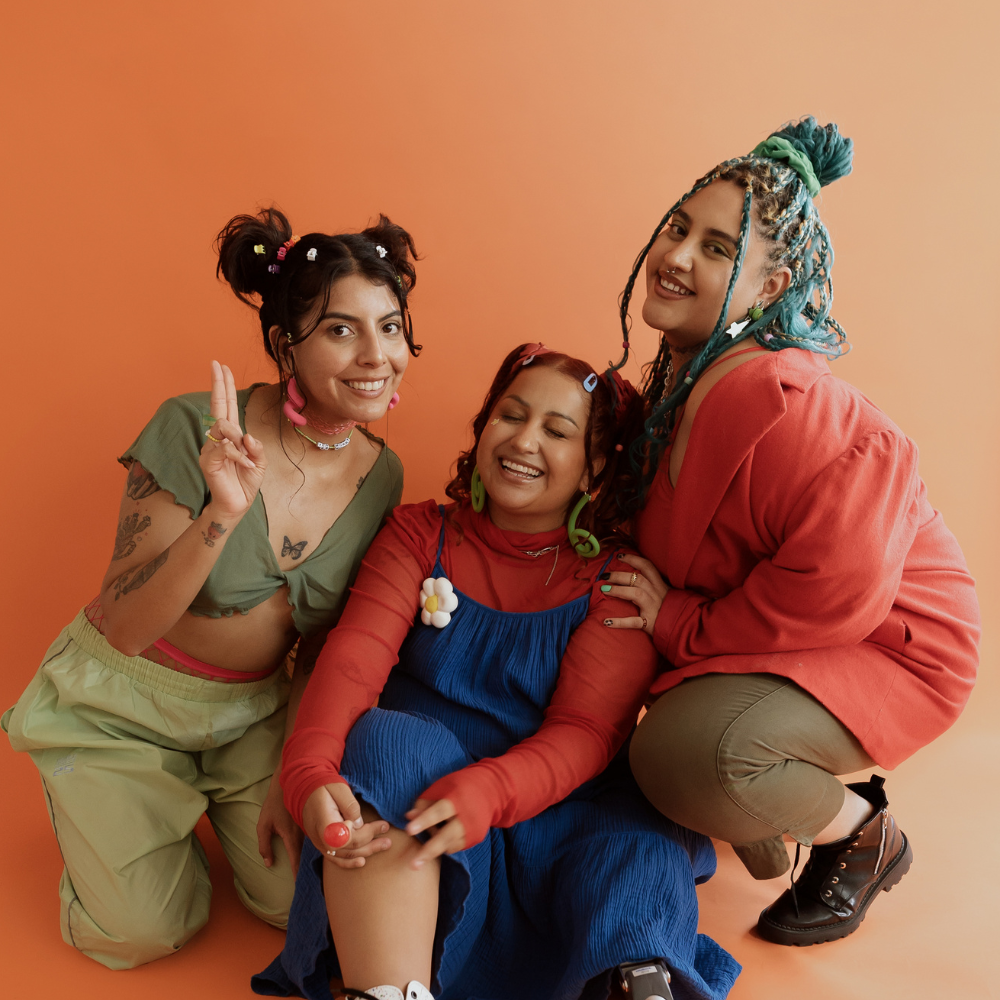 Three women with colorful hairstyles and vibrant outfits posing together against an orange background, showing happy and playful expressions.