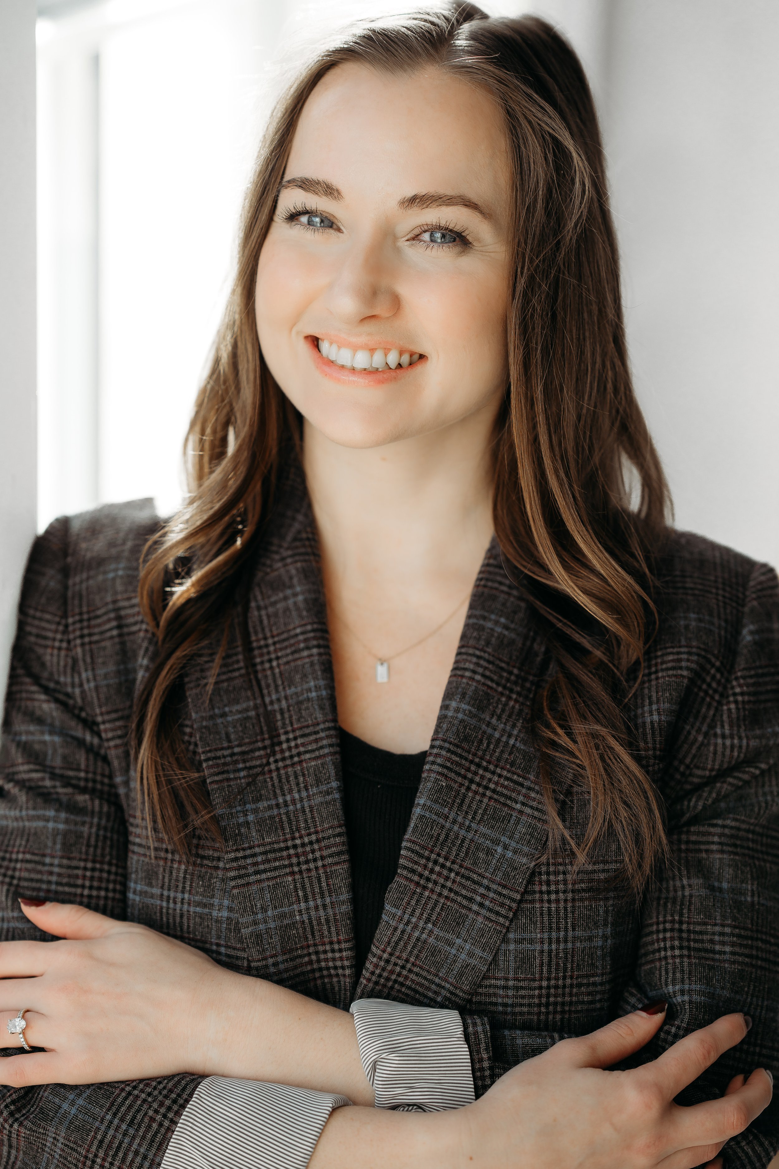 Portrait of a woman with long brown hair, blue eyes, smiling, wearing a dark plaid blazer, a black top, a silver necklace, and a ring on her left hand, standing near a window.
