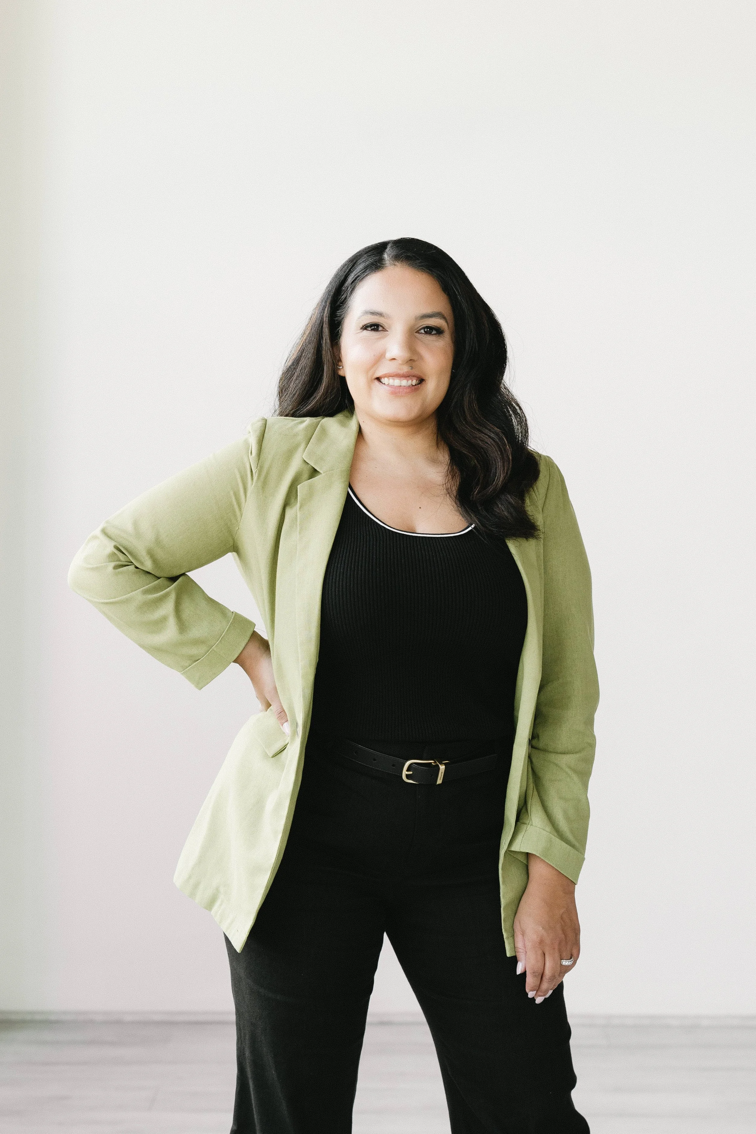 Woman with long dark hair wearing a light green blazer and black top, standing against a plain white wall.