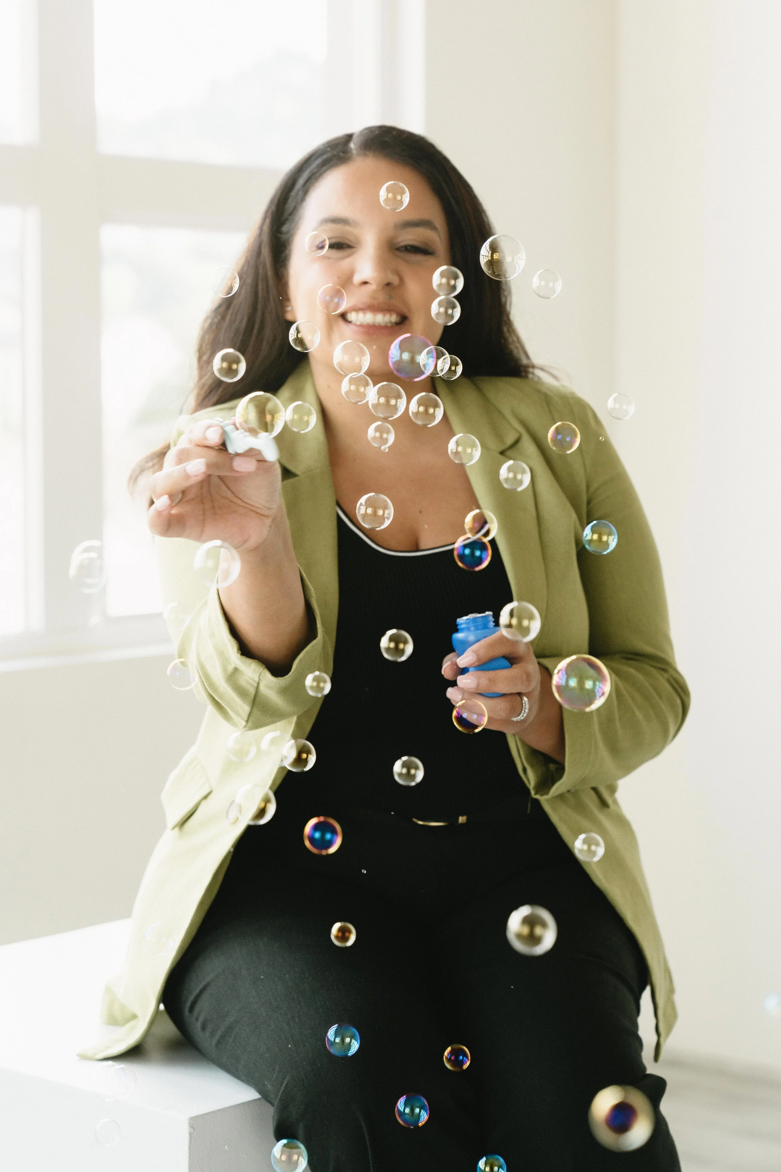 A woman in a yellow blazer and black shirt sitting near a window, blowing soap bubbles.