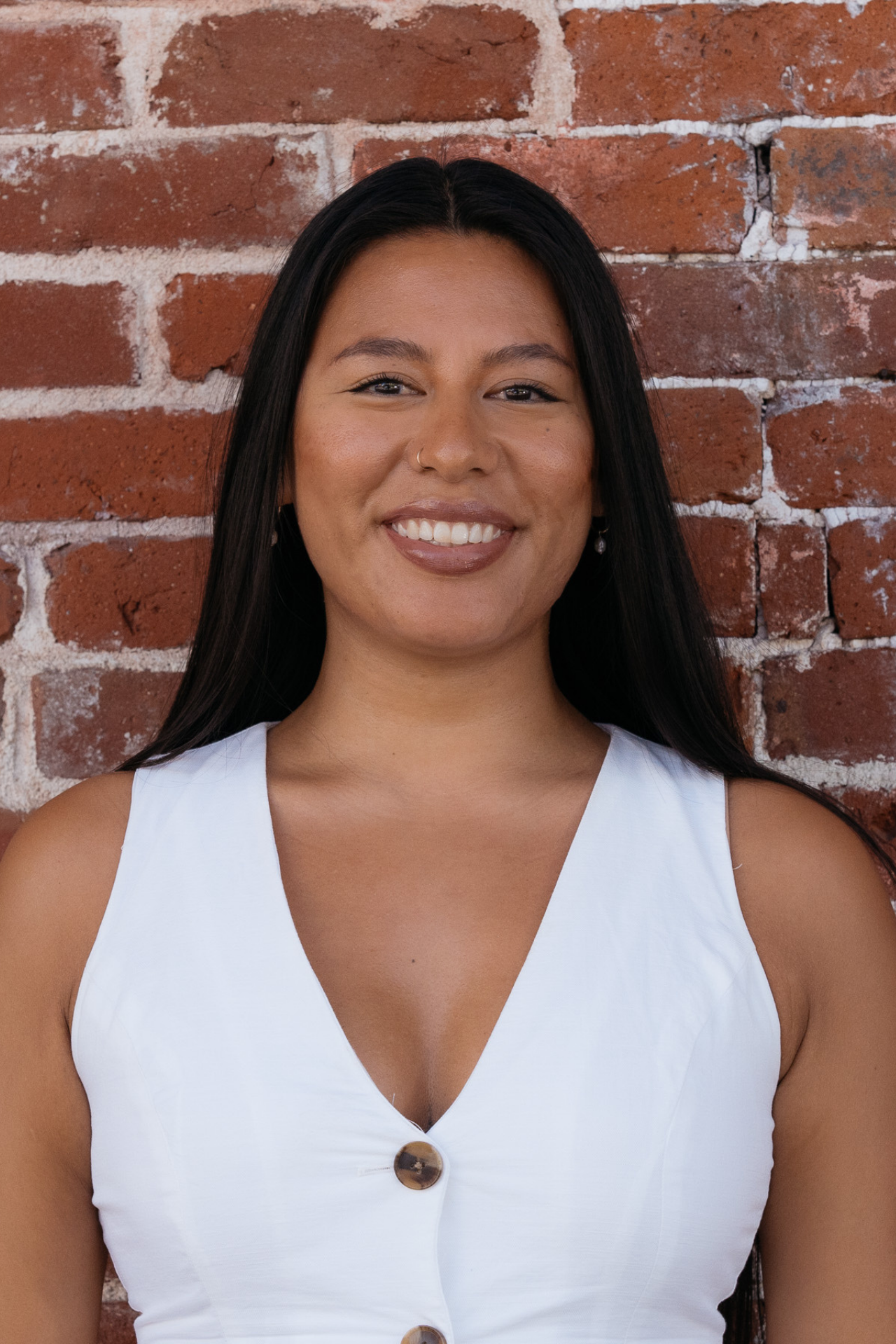 A smiling woman with long dark hair and brown skin standing in front of a red brick wall, wearing a sleeveless white top with buttons.