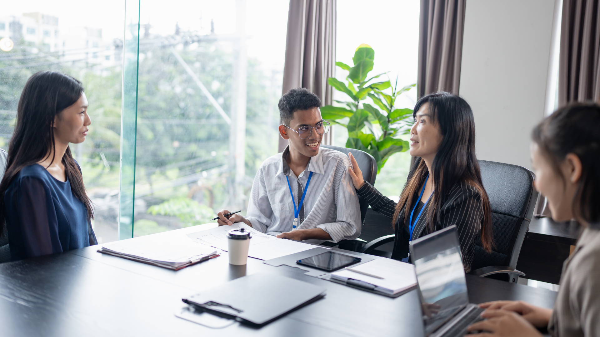 Five diverse people in a business meeting room engaged in conversation, sitting around a table with laptops, documents, and a coffee cup, with large windows and a green plant in the background.