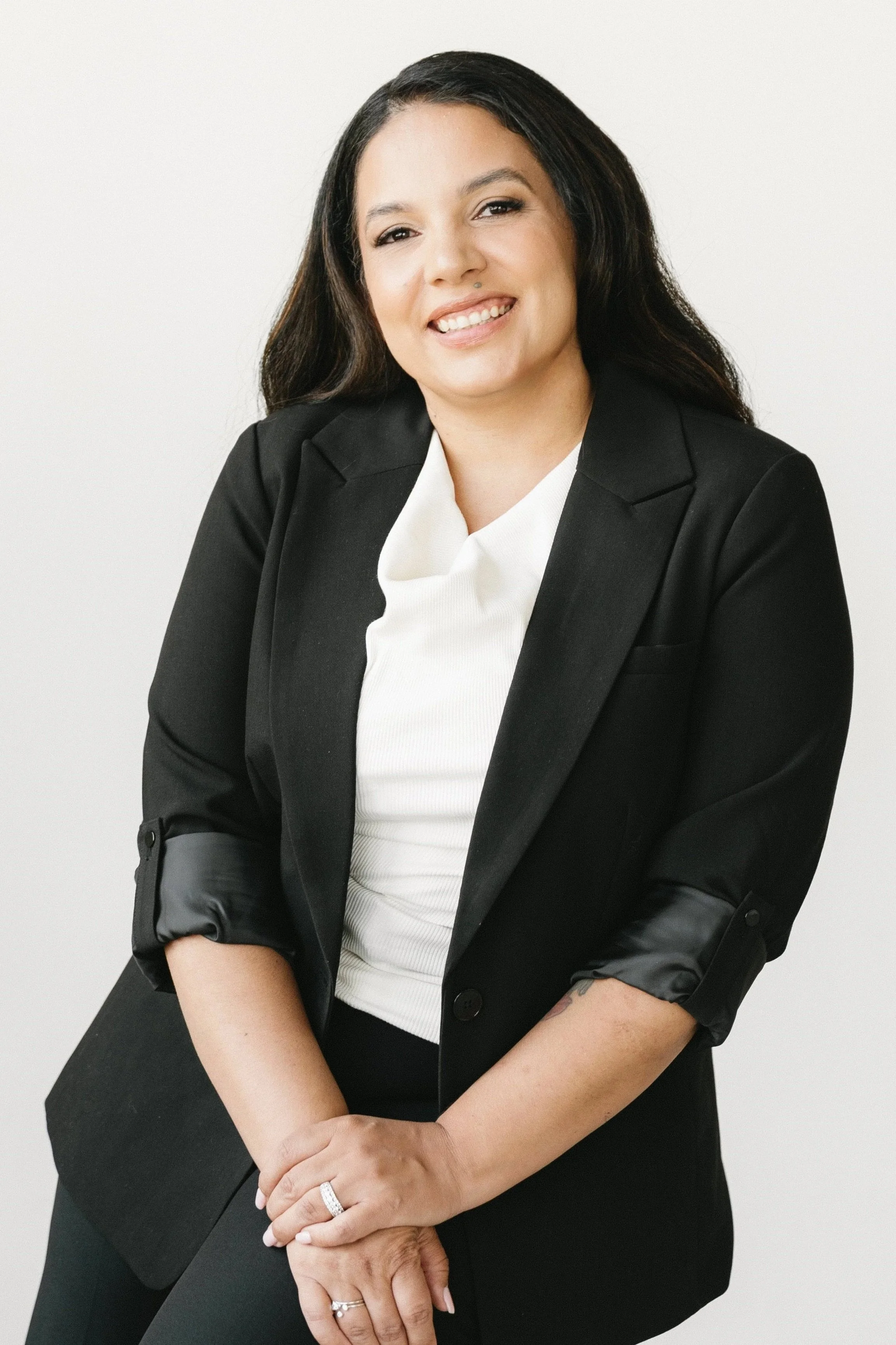 A woman with dark hair and a light skin tone wearing a black blazer and white top, smiling, with her hands clasped in front, against a plain white background.