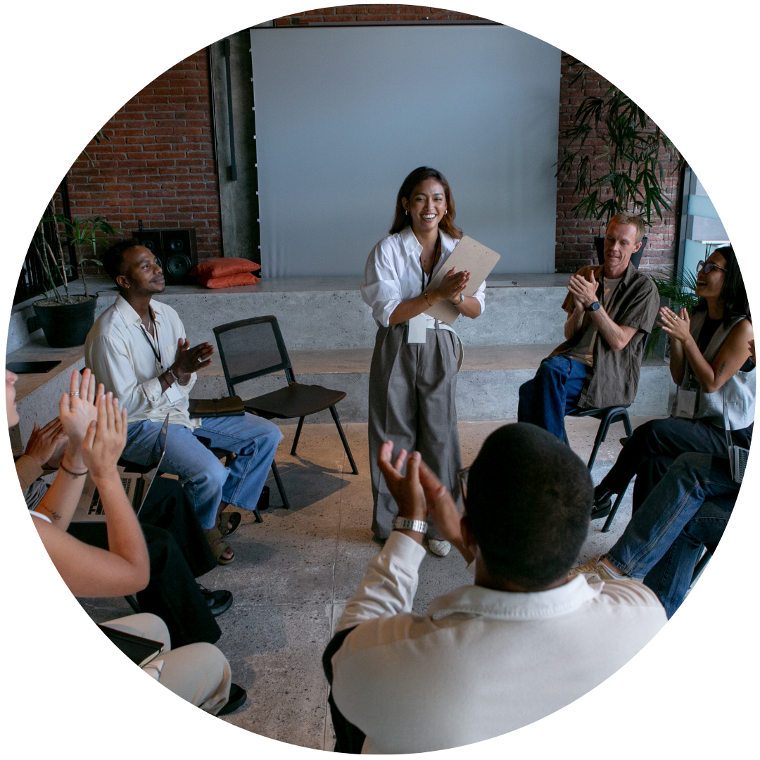 A diverse group of people clapping and smiling as a woman stands in the center holding a clipboard, leading a discussion or presentation in a modern, casual workspace.