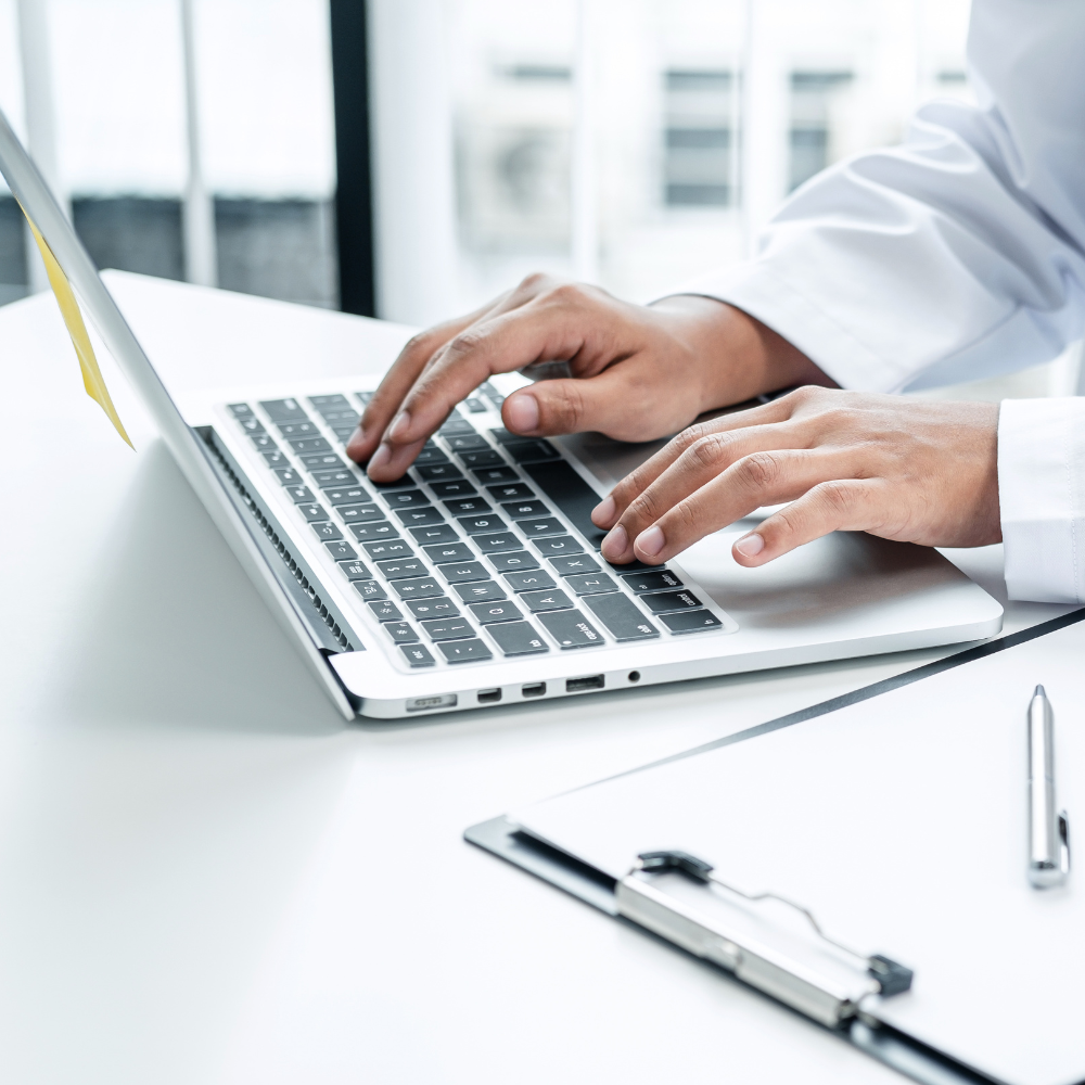 Person typing on a laptop keyboard at a white desk, with a clipboard and pen nearby in a bright office setting.