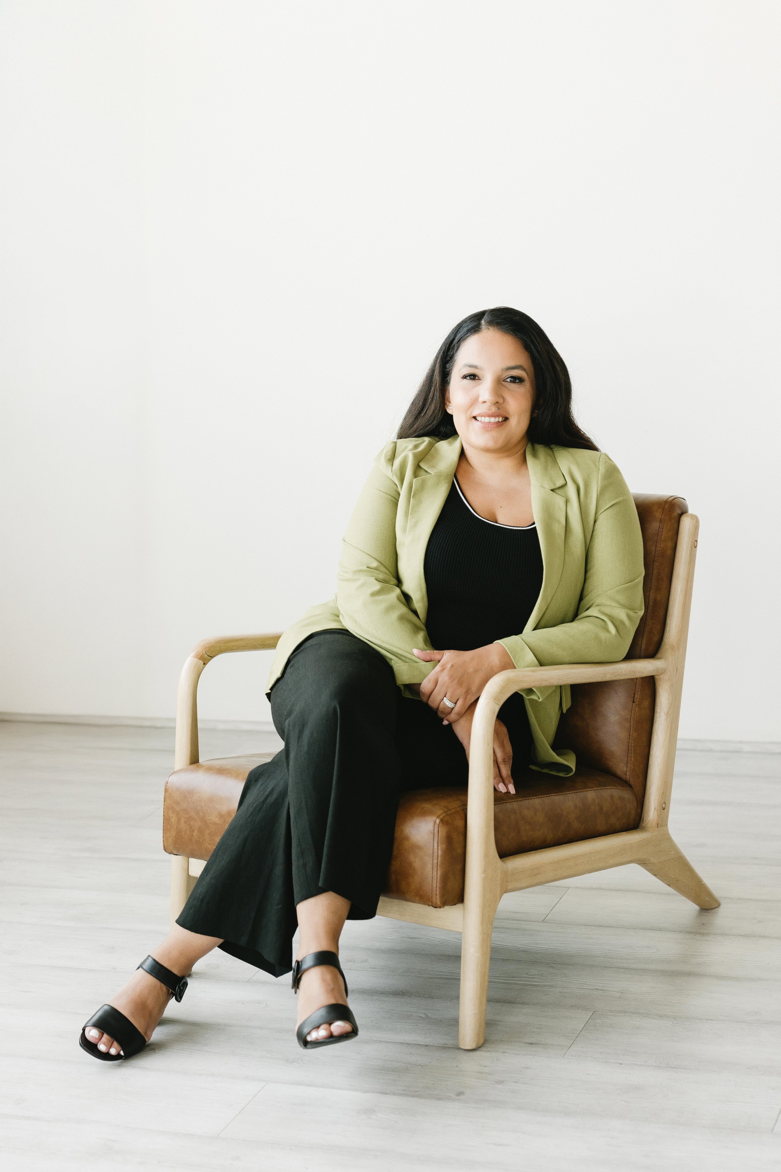 Woman sitting on a wooden chair with leather cushion, wearing a green blazer, black top, black pants, and black sandals, smiling indoors against a white wall.