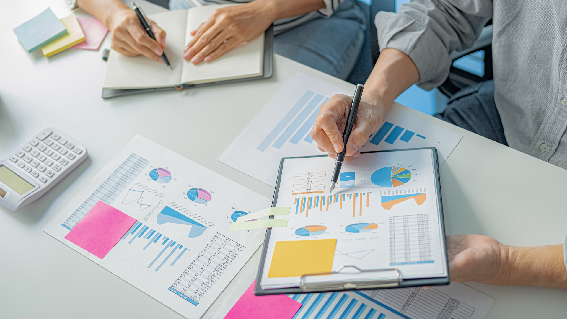 People working on data analysis with charts and graphs on paper and a tablet, on a white desk with sticky notes, calculator, and notepad.