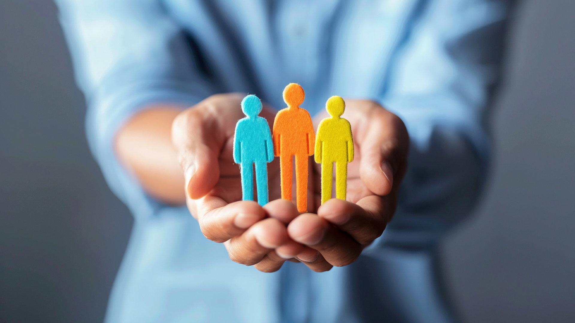 A person's hand holding three colorful felt figures representing people in blue, orange, and yellow, with a gray background.