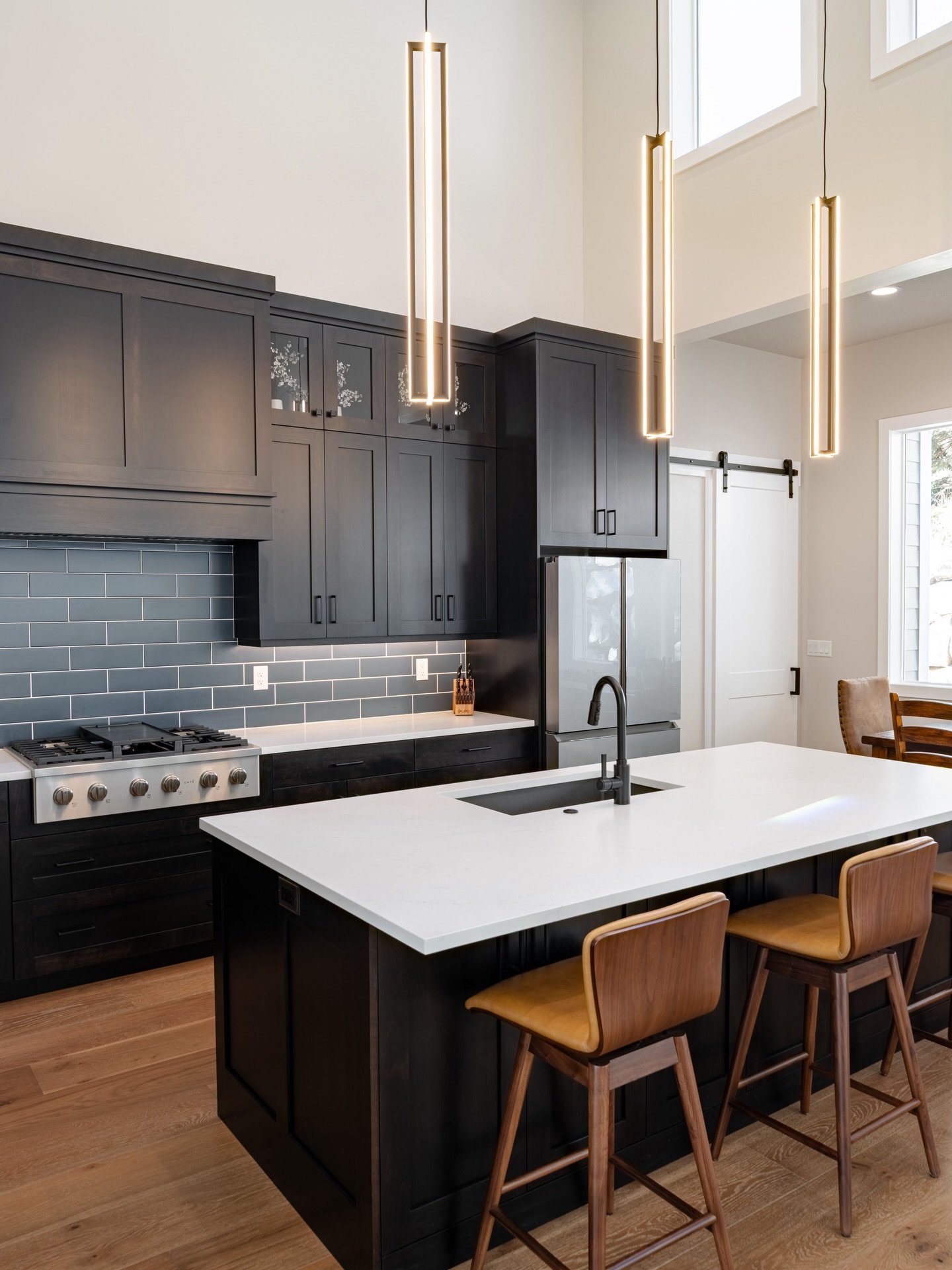 Double-height only works if it&rsquo;s grounded. The floor-to-ceiling cabinetry gives the space weight, while the darker tile anchors the kitchen to where you spend your time. Linear pendants pull the volume back down so the room feels dramatic, but 