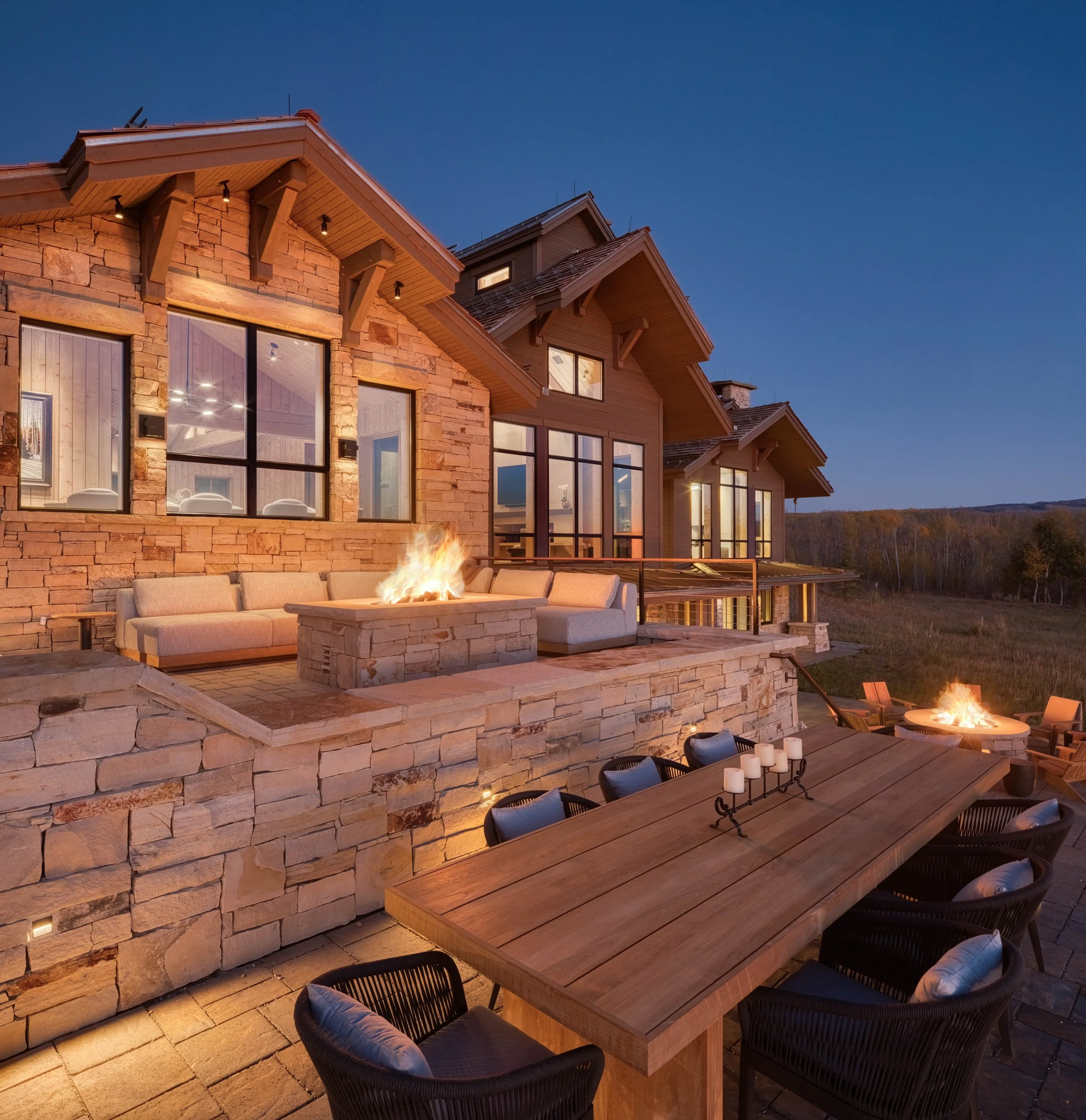 A spacious outdoor patio at dusk with stone and wood house in the background, fireplace, seating area, and a dining table with chairs.