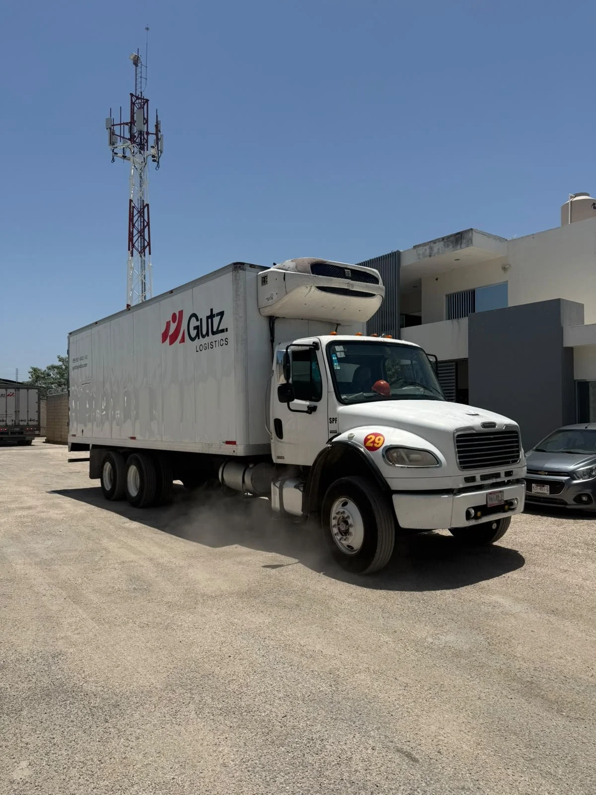 Camión de logística blanco con el logo de Gutz Logistics, equipado con refrigeración, en un área de estacionamiento con edificios modernos y un fondo de cielo despejado.