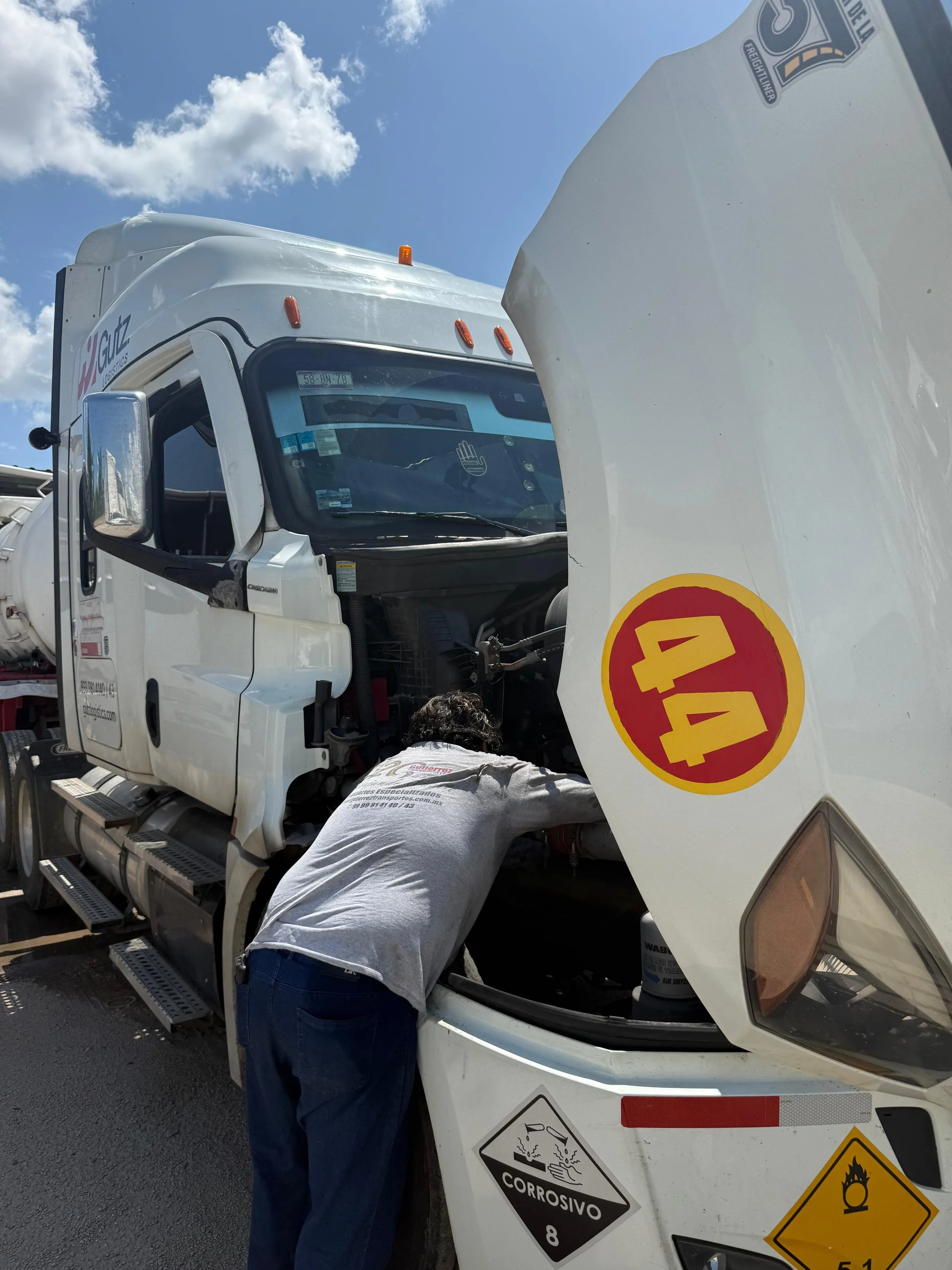 Persona trabajando en el motor de un camión de carga blanca, con señales de advertencia de peligro químico, corrosivo y peligro de explosión.