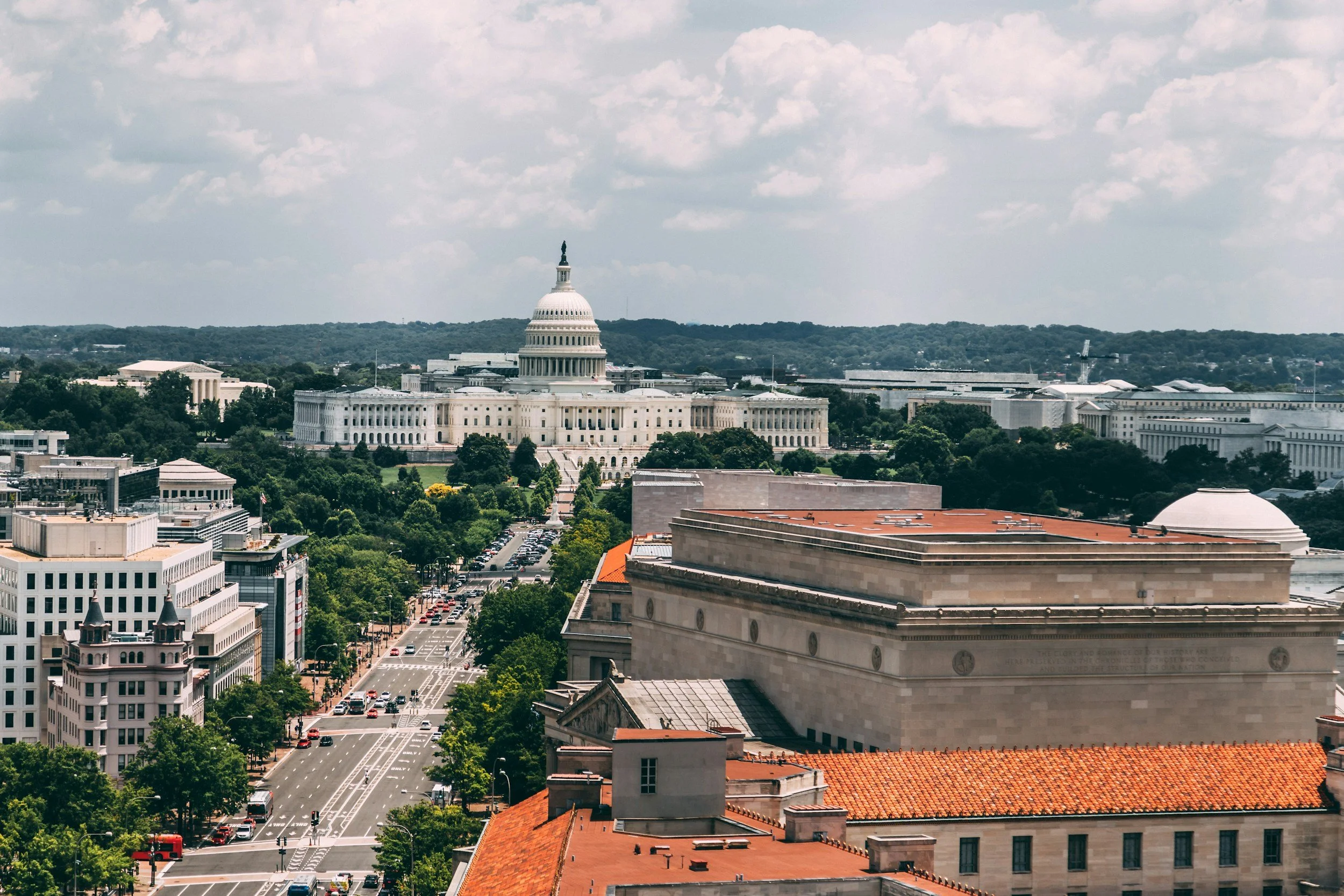 A cityscape view of Washington D.C., featuring the United States Capitol building with its iconic dome in the background, surrounded by various government and office buildings, green trees, and a busy street with cars.