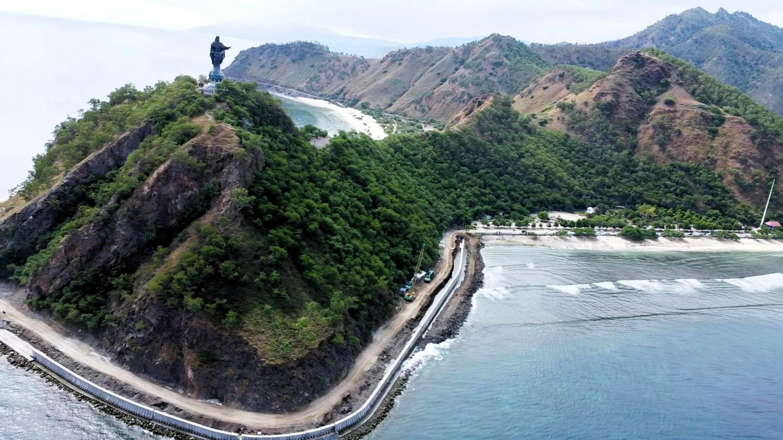 A coastal hill in Dili, Timor-Leste with a statue at the top, surrounded by dense green trees and a lookout road running along the edge, overlooking the ocean.