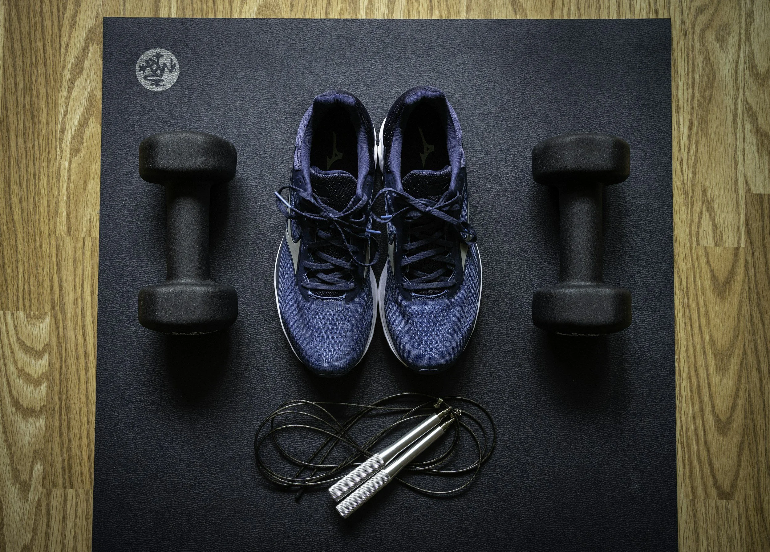blue running shoes flanked by dumbells  and skip rope over a yoga/exercise mat with a little of the wooden floors underneath showing