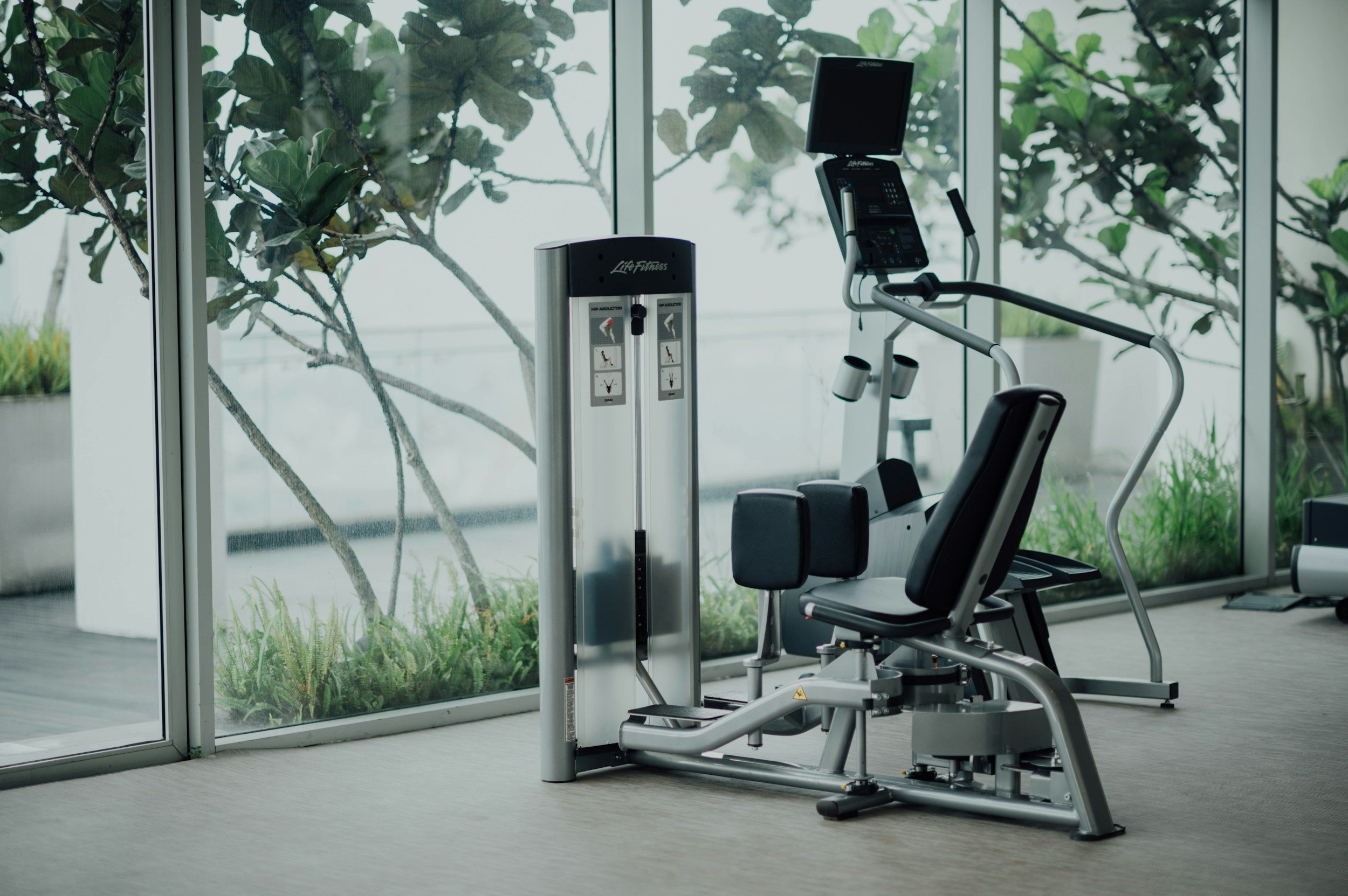 Empty fitness gym with a seated leg press machine and an elliptical trainer, large floor-to-ceiling windows with a view of indoor plants and trees outside.