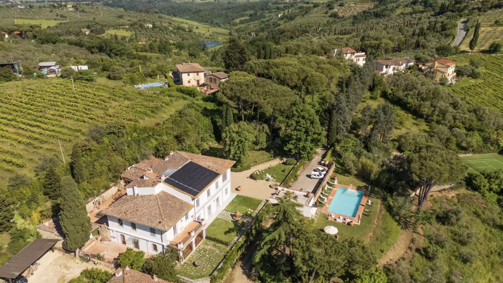 Aerial view of a countryside estate with main house, swimming pool, and surrounding lush green landscape, hills, vineyards, and scattered houses in the background.