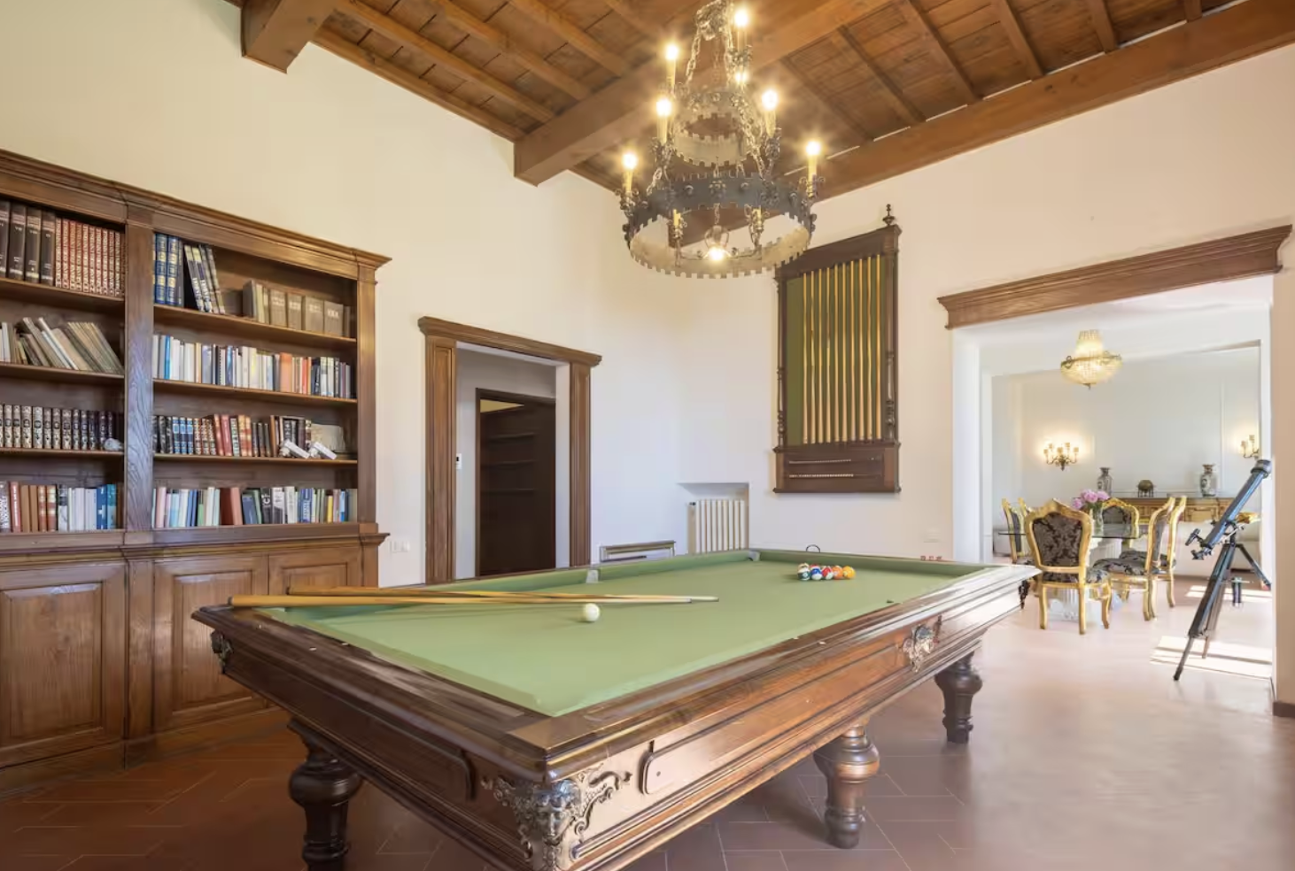 Elegant room with a wooden pool table, a bookshelf filled with books, and a chandelier hanging from the wooden ceiling. Adjacent to the pool table is a dining area with ornate chairs and a chandelier.