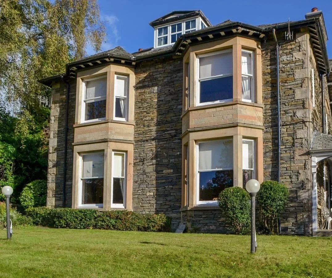 A multi-story house with a stone facade and large bay windows, situated on a well-maintained lawn with two white globe street lamps and bushes, under a clear blue sky.