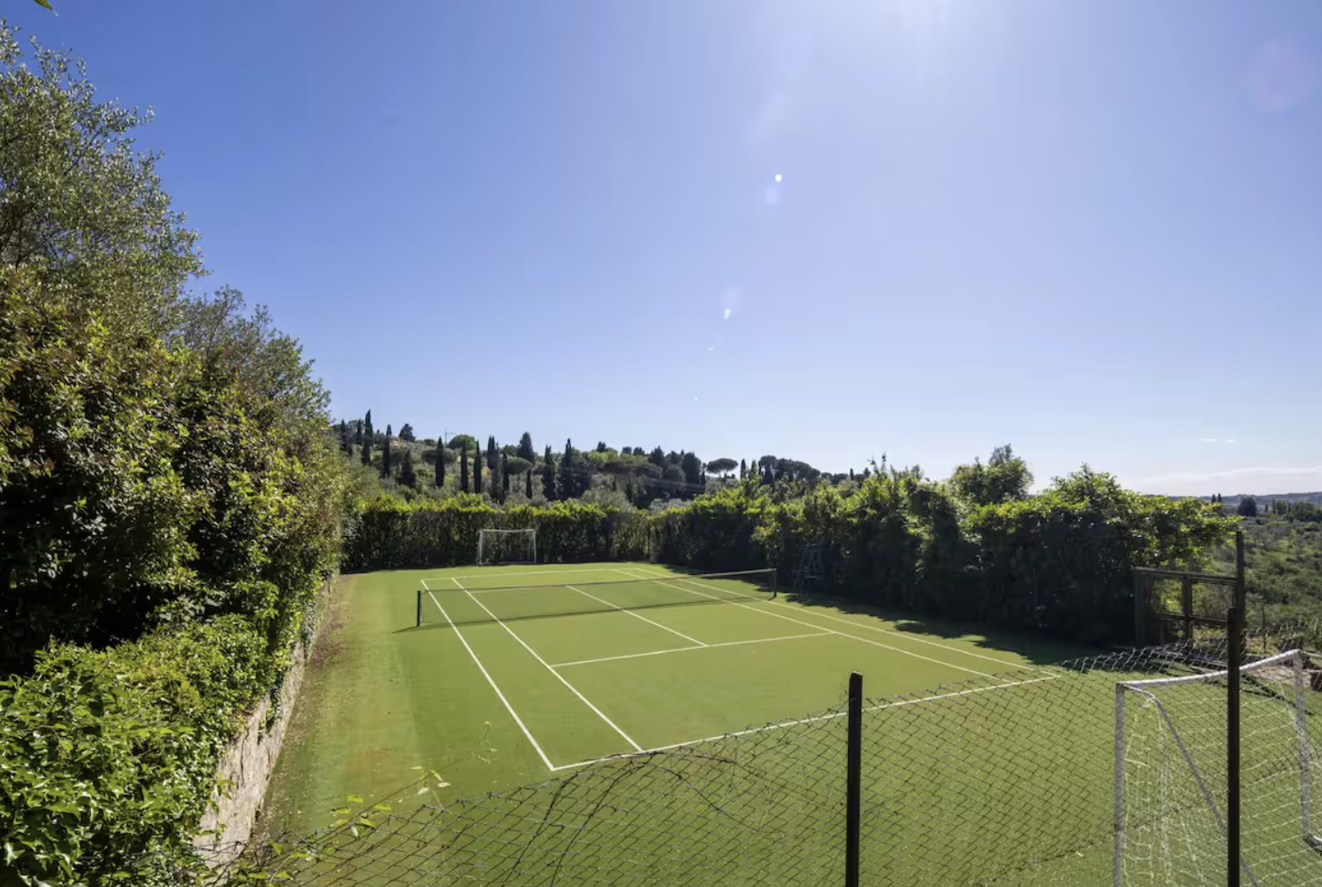 Empty tennis court surrounded by green hedges and trees under a clear blue sky.