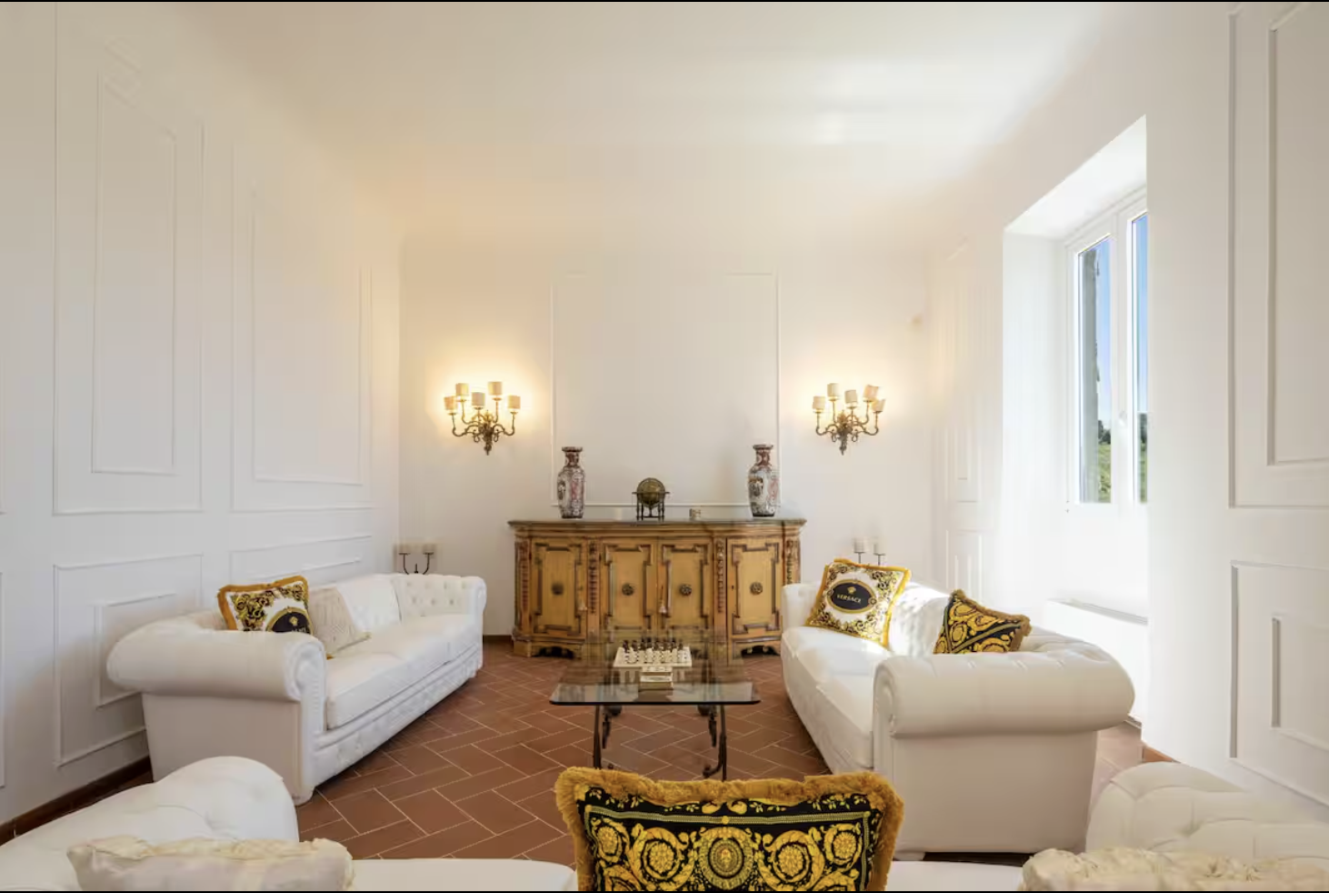 Bright living room with white sofas, a glass coffee table, a wooden sideboard, wall lamps, decorative pillows, and a large window allowing natural light.