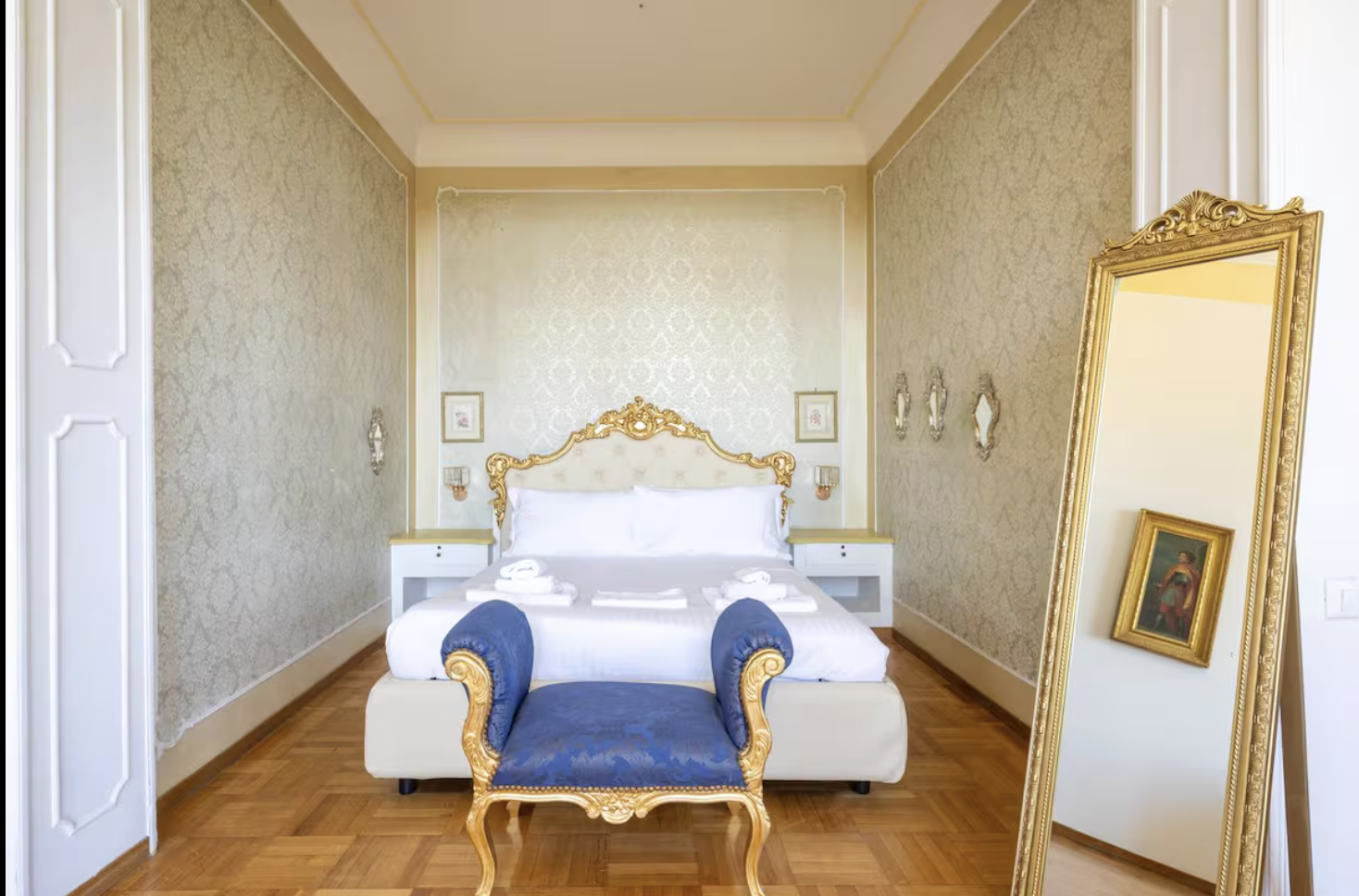 Luxury bedroom with ornate gold and blue furniture, a mirror and picture frame, and a neatly made bed with towels.