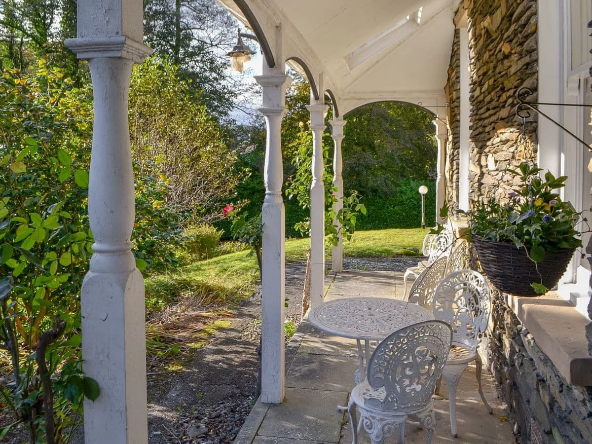 A porch with white decorative iron chairs, a small round table, hanging flower basket, and garden with bushes and trees in the background.