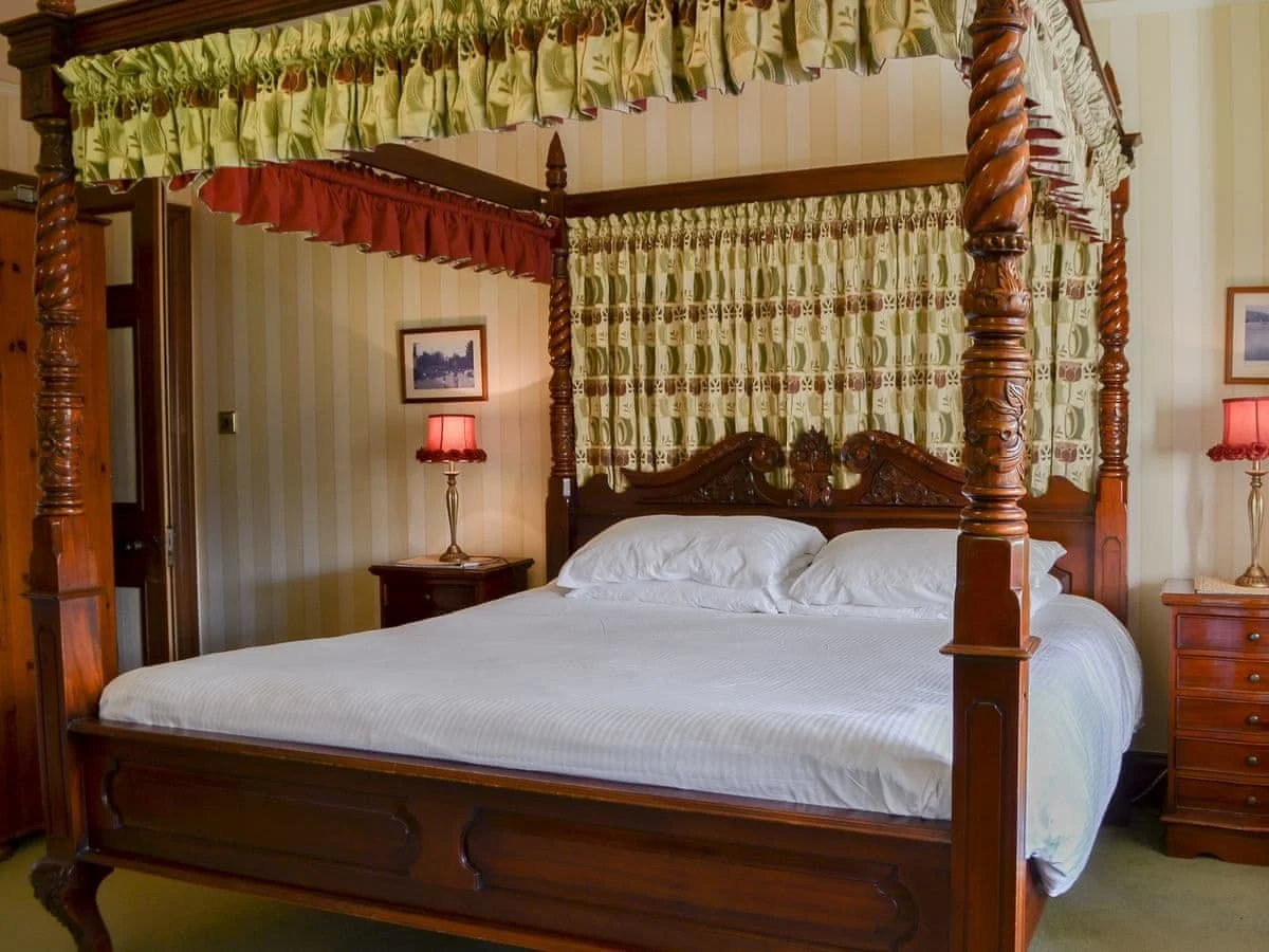 A traditional four-poster bed with wooden carvings, white bedding, and a canopy with patterned curtains in a vintage bedroom.