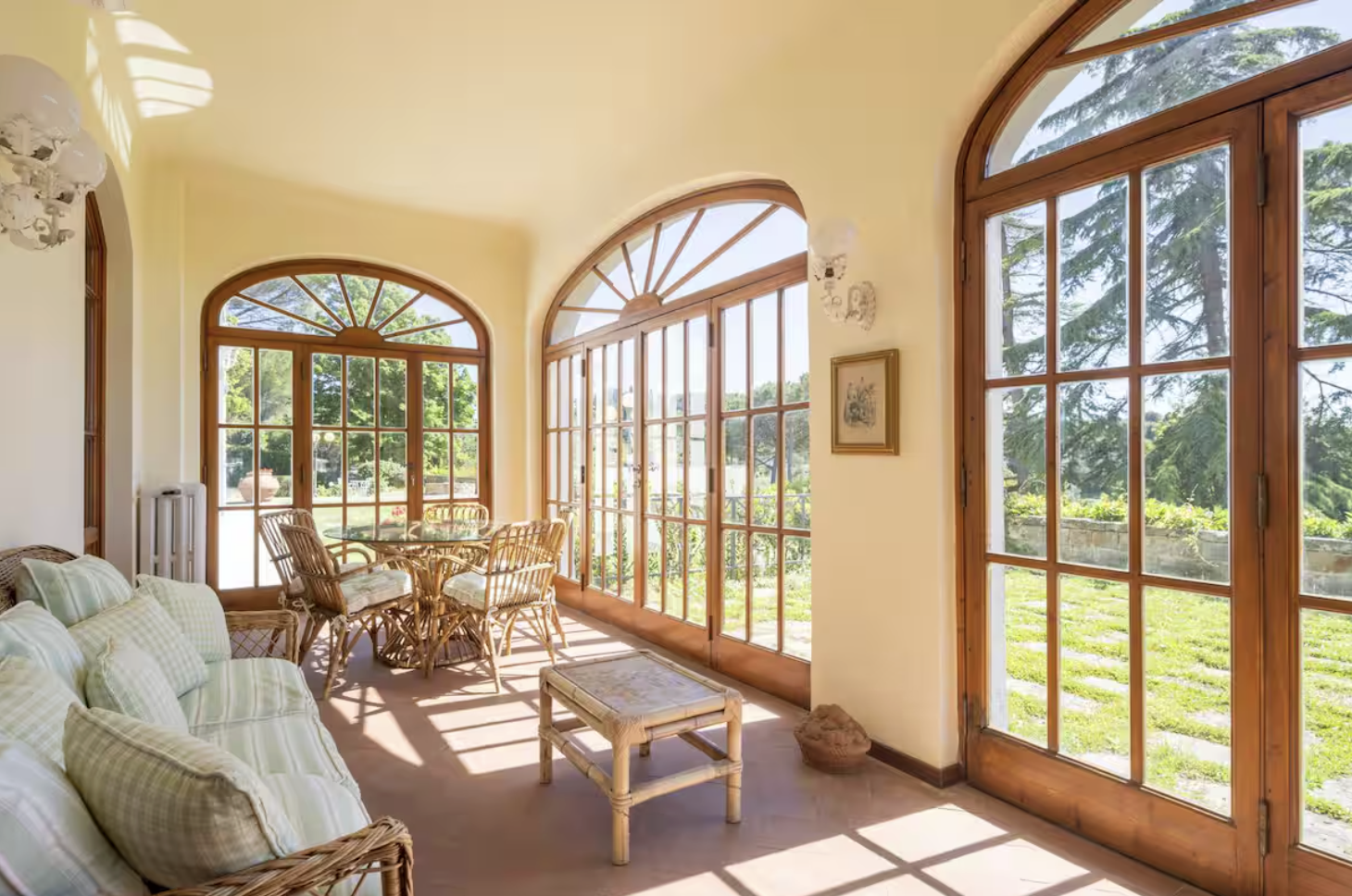 Bright sunlit sunroom with large arched windows and glass doors, wicker furniture, a cushioned sofa, a round dining table with wicker chairs, and a view of greenery outside.