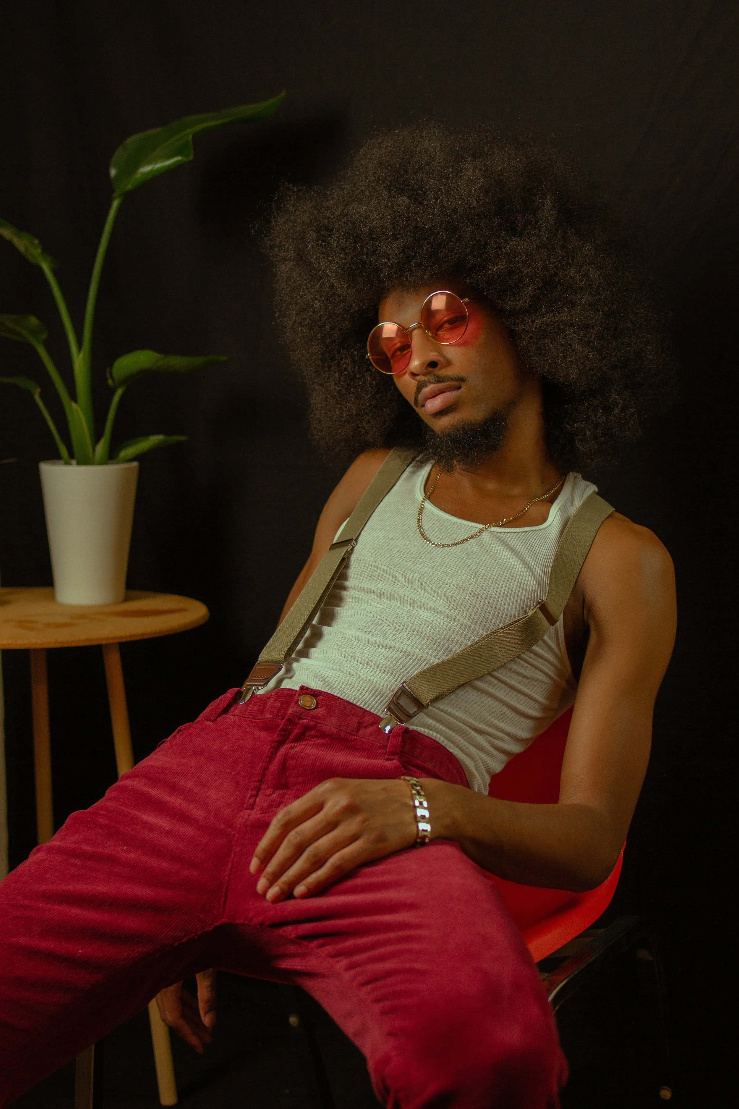 A man with an afro hairstyle wearing pink-tinted sunglasses, a beige tank top, red pants, suspenders, a necklace, and a bracelet, sitting in a red chair against a black background with a small wooden table and a potted plant nearby.
