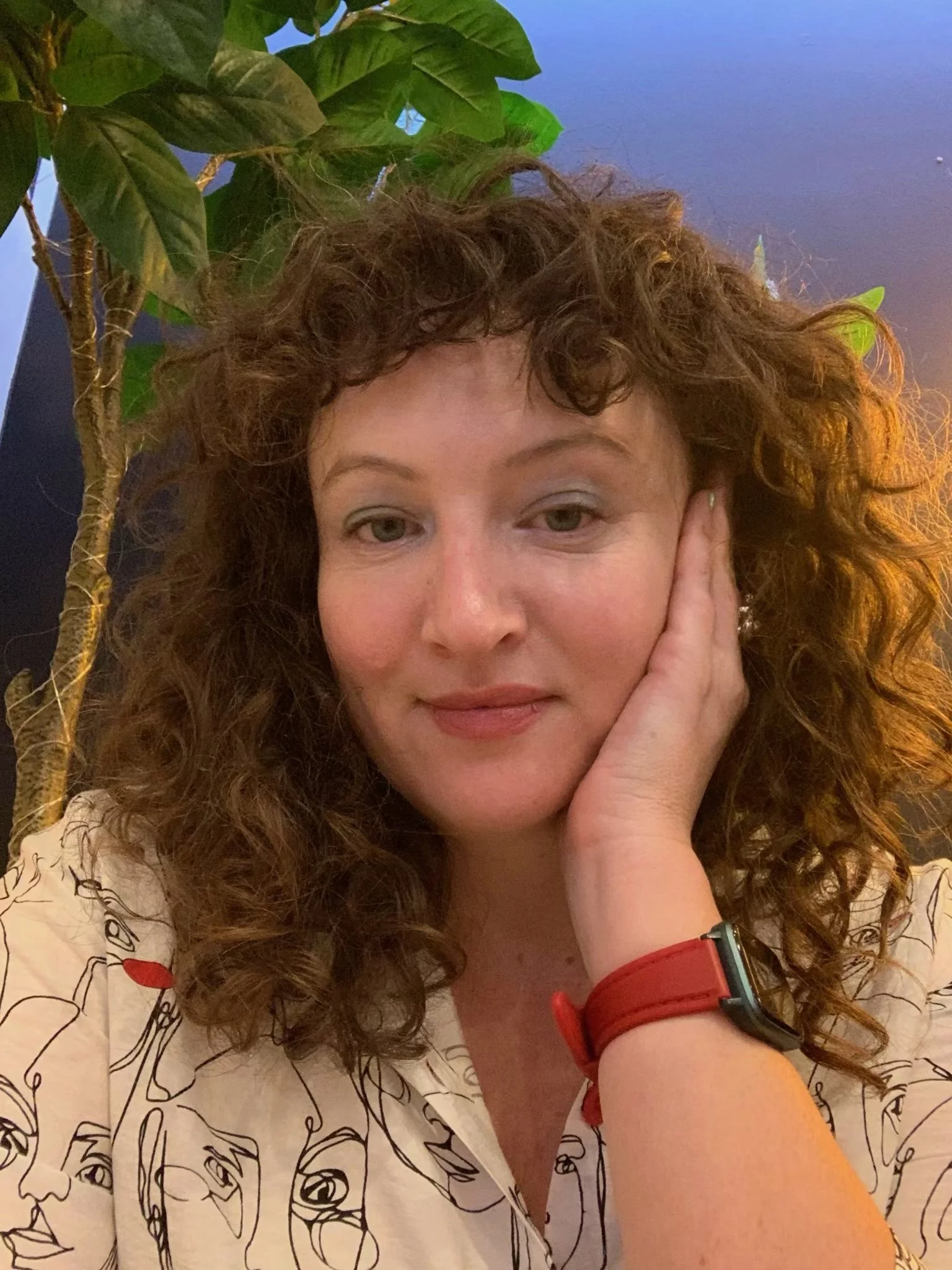 A woman with curly brown hair, wearing a white shirt with abstract face sketches, resting her head on her hand, in front of a plant with large green leaves.
