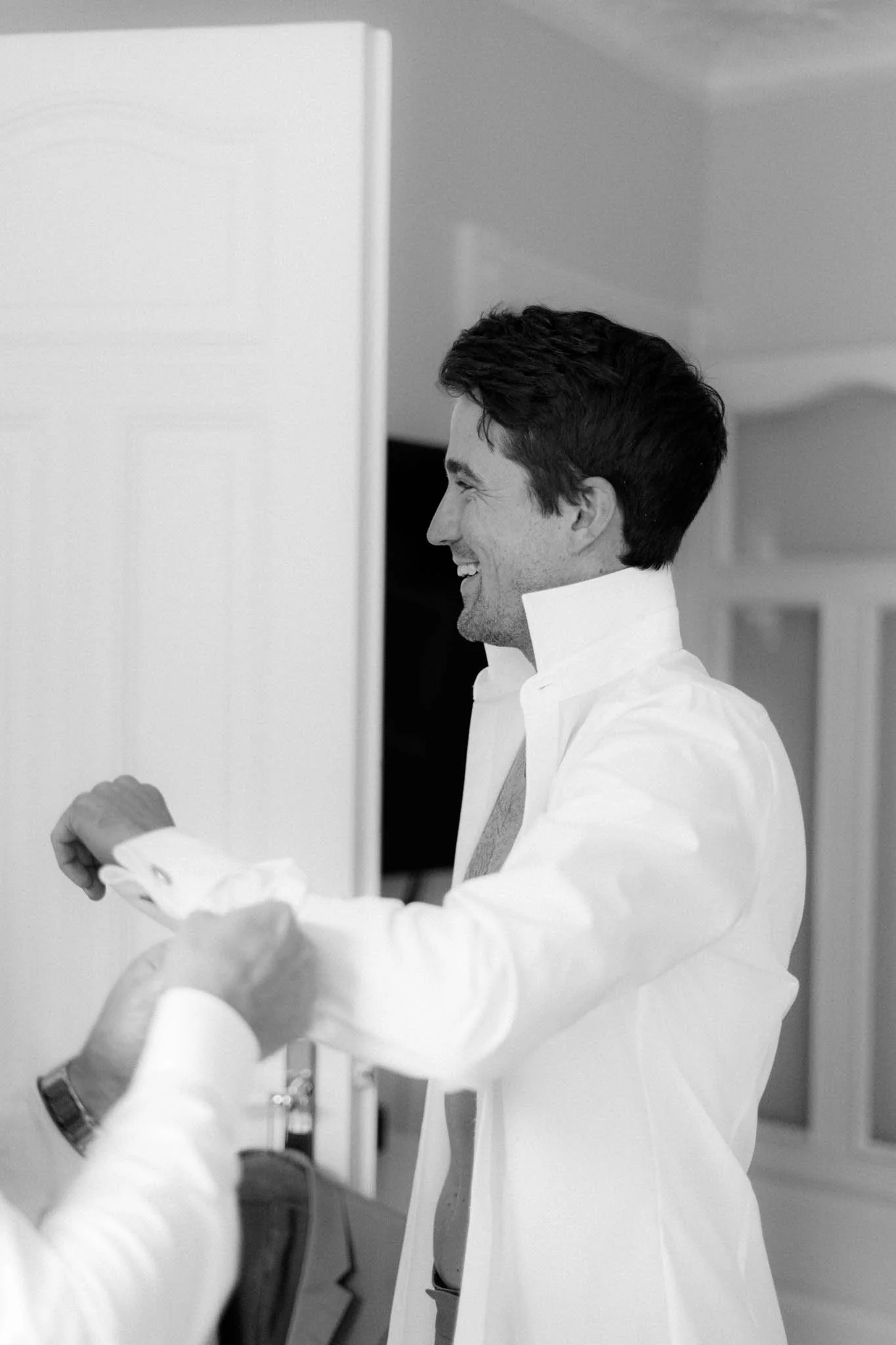 Ein lachender Mann in einem weißen Hemd beim Getting Ready bekommt Hilfe beim Zuknöpfen der Ärmel von seinem Trauzeugen.