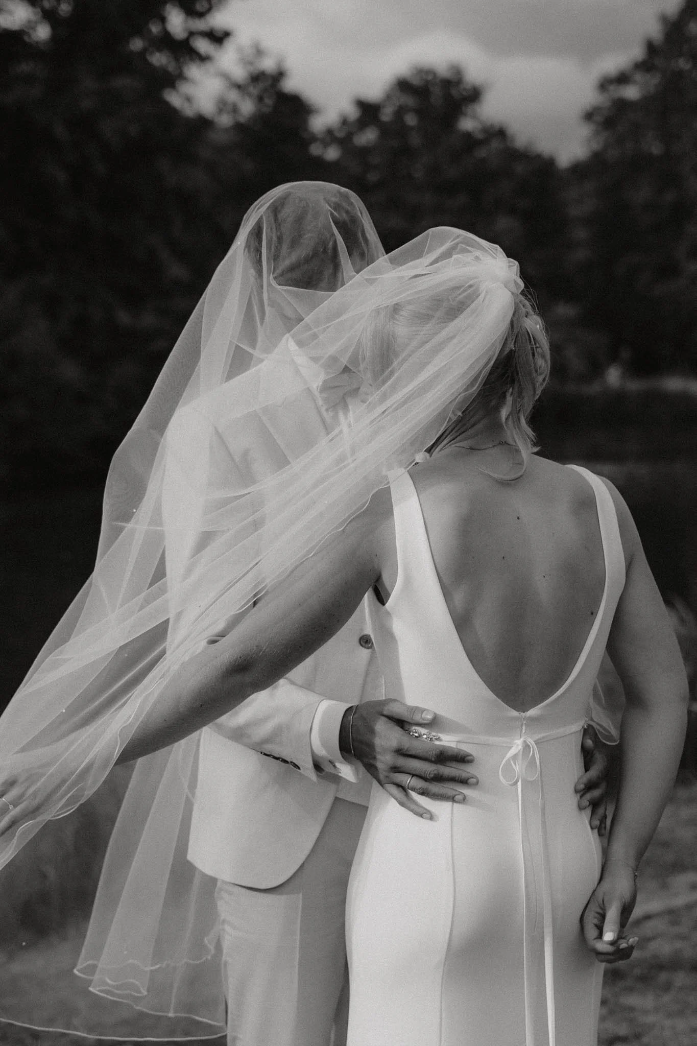 Zwei Menschen bei einem Hochzeitsfoto, die sich umarmen, mit einer Braut in einem weißen Kleid und einem Schleier, im Freien mit Bäumen im Hintergrund