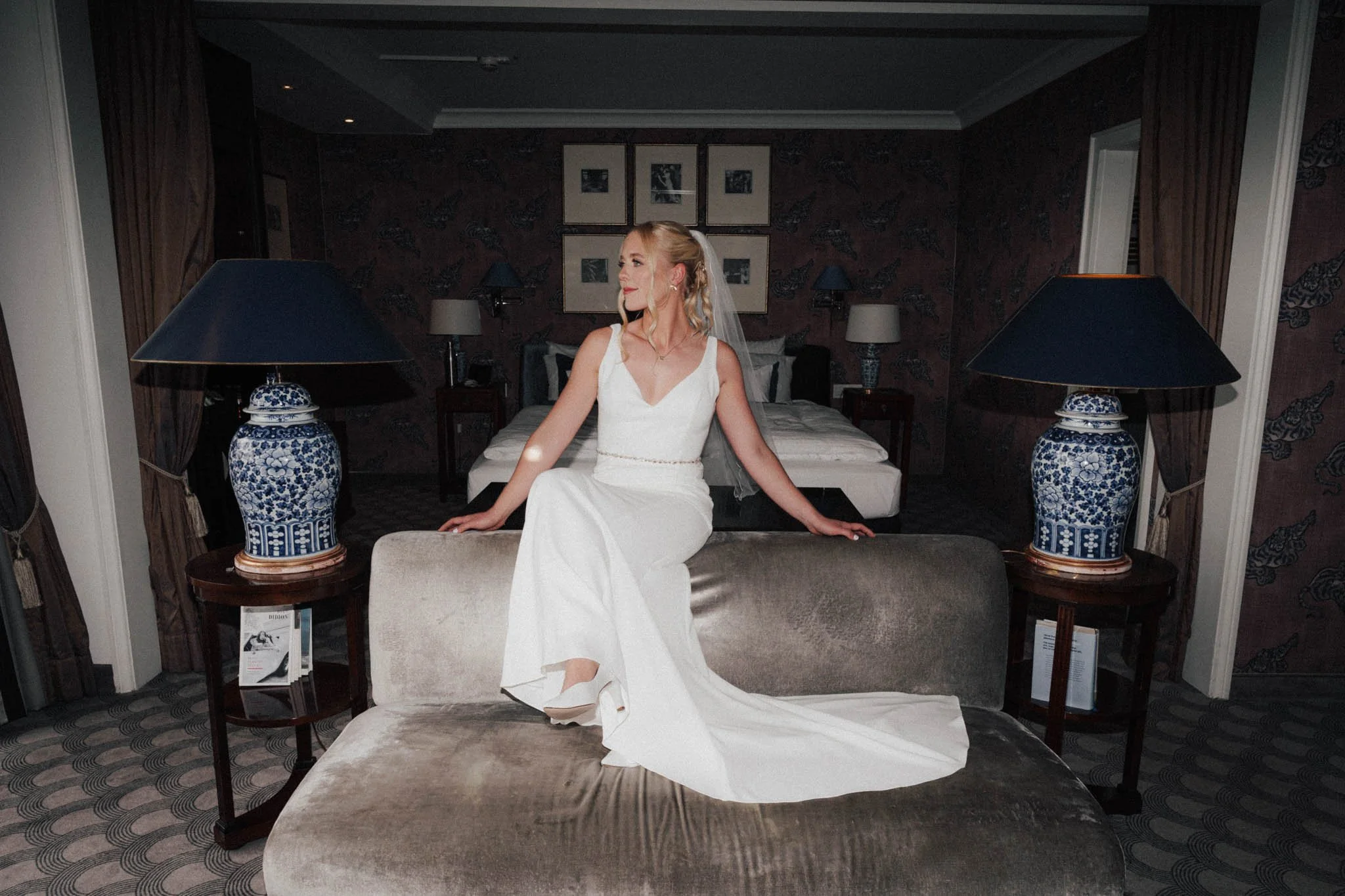 Braut in weißem Hochzeitskleid sitzt auf einer Couch in einem Hotelzimmer, im Hintergrund Bett und Wandbilder.