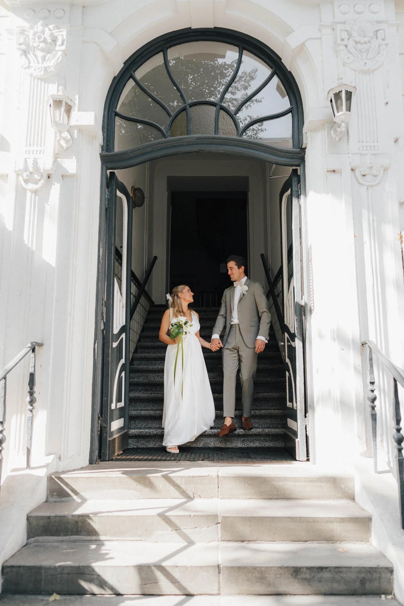 Braut und Bräutigam gehen die Treppe eines weißen Gebäudes (Altbau in Hamburg) hinunter, halten Händchen und lachen, die Braut trägt ein weißes Kleid und die Blume, der Bräutigam einen grünlichen Anzug.