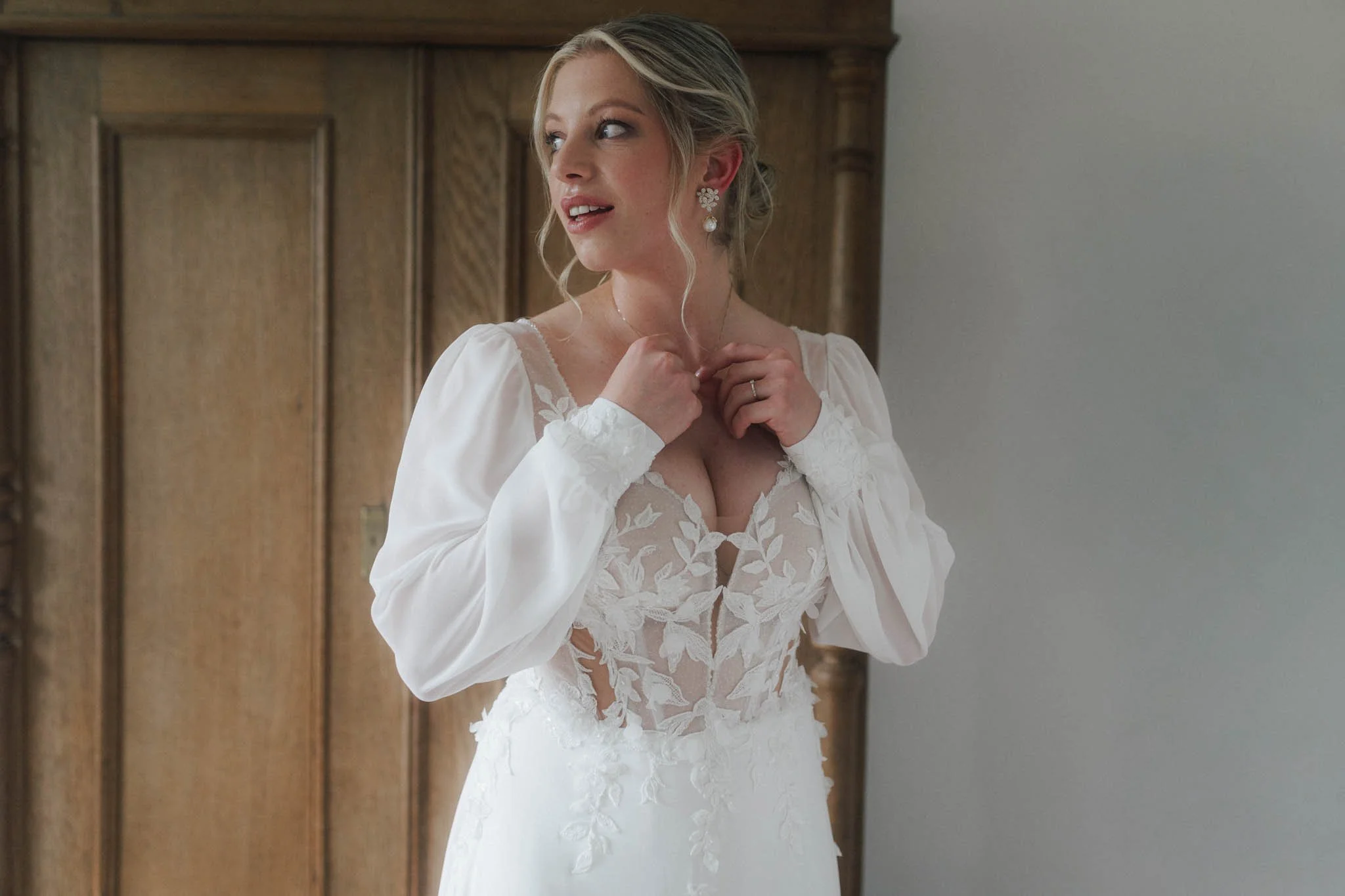 Junge Braut in weißem Brautkleid mit Spitzenmuster, die im Zimmer steht, ihre Haare zurechtmacht und sich Freude zeigt.