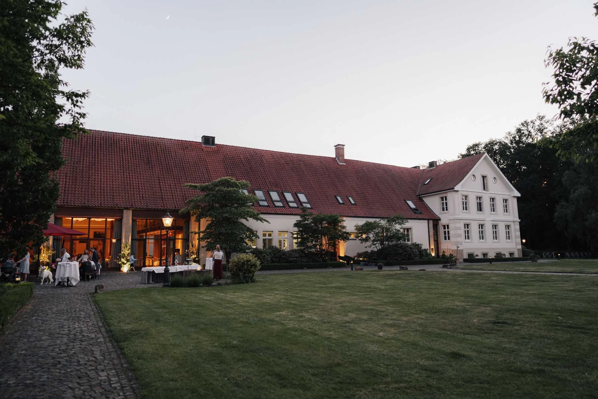 Ein historisches weißes Gebäude mit rotem Ziegeldach bei Dämmerung, umgeben von einem gepflegten Garten mit Blumen und Bäumen, mit Gästen, die auf der Terrasse Feierlichkeiten im Freien genießen.