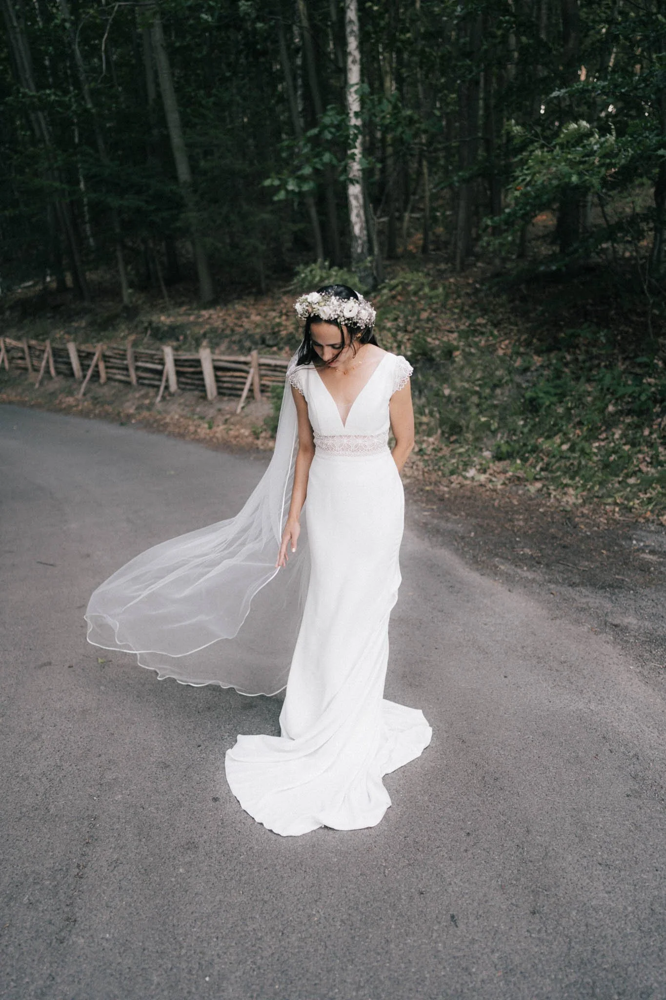 Eine Frau in einem weißen Hochzeitskleid mit Blumenkranz auf dem Kopf steht auf einer Straße vor einem grünen Wald.